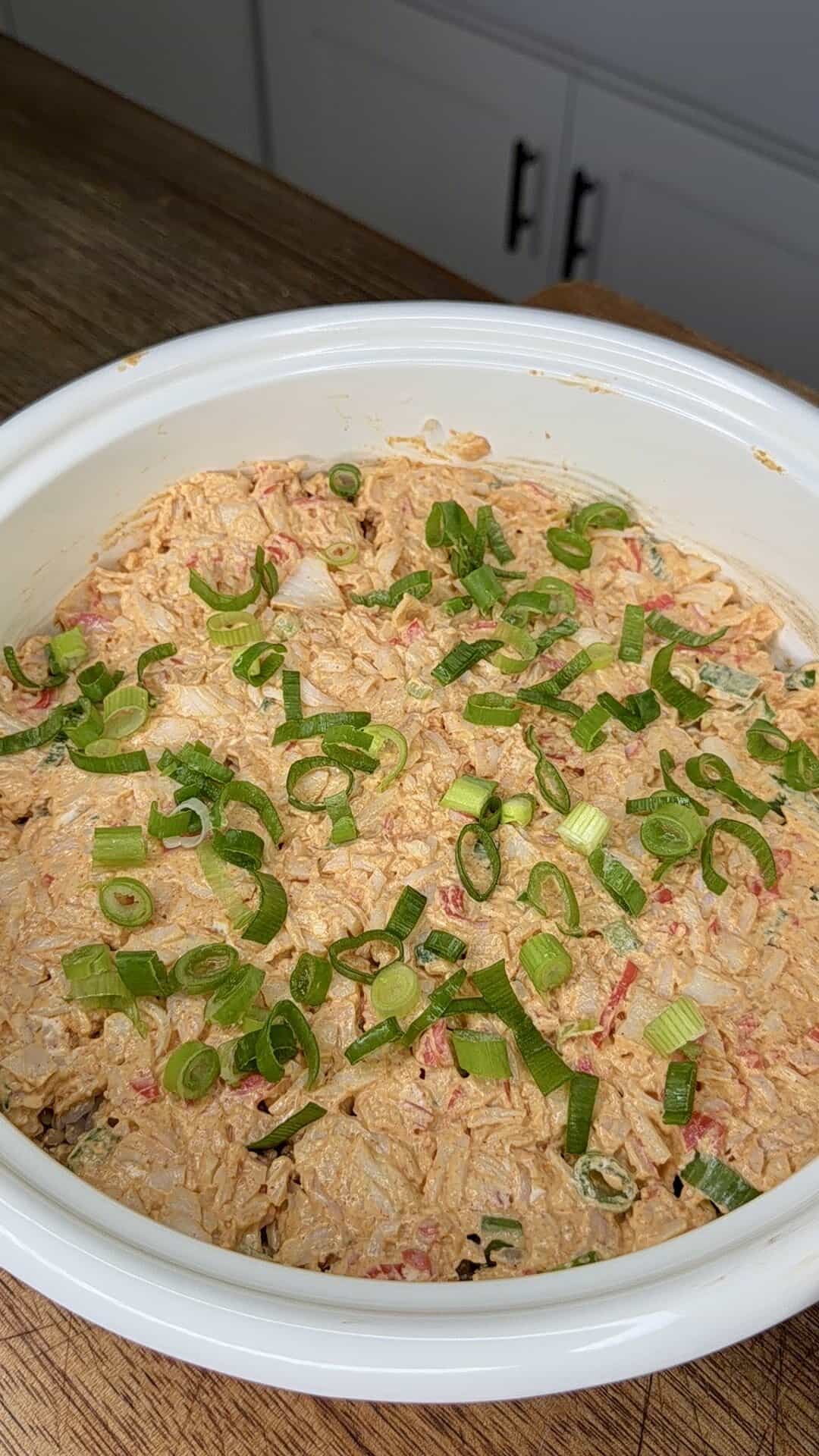 A white dish filled with a creamy, shredded chicken mixture topped with sliced green onions, resting on a wooden surface in a kitchen.