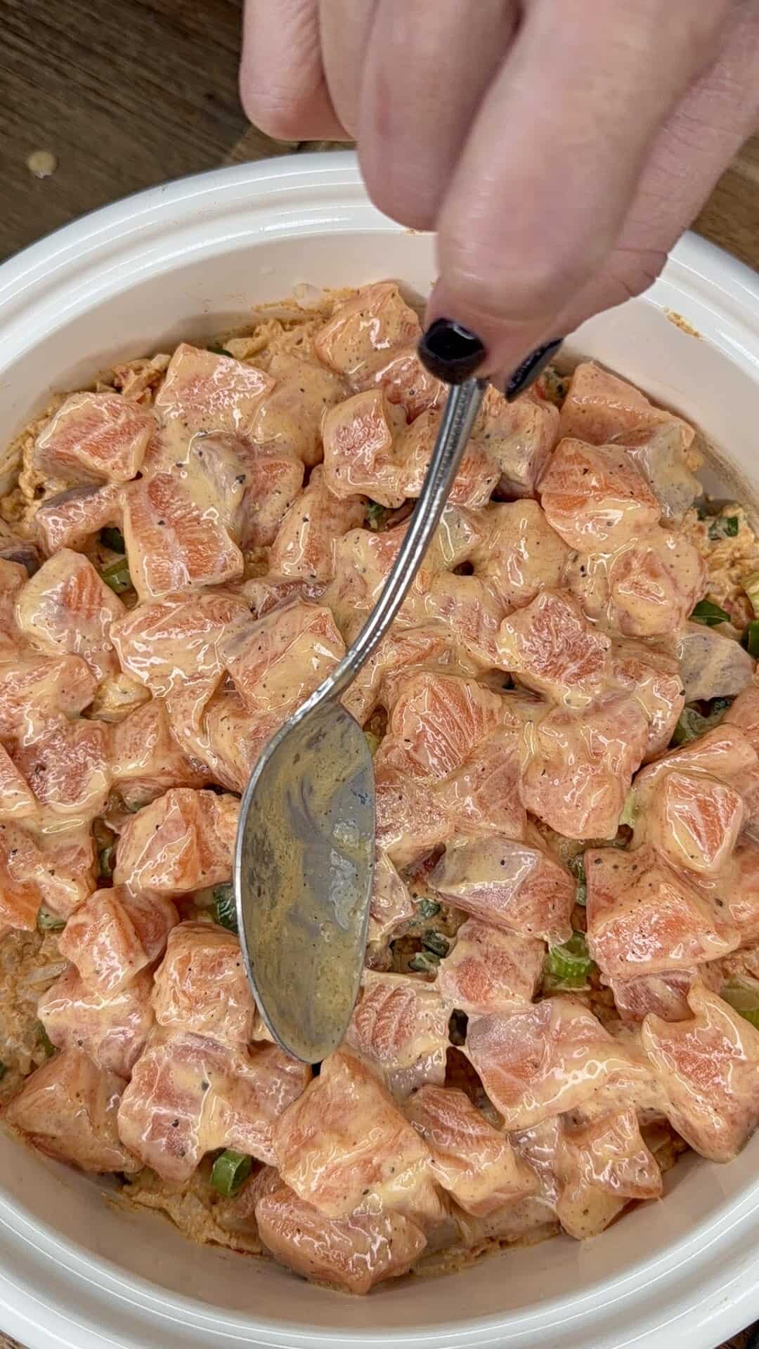 A hand with dark nail polish holds a spoon, mixing chunks of raw salmon coated in a creamy sauce with herbs in a white bowl.