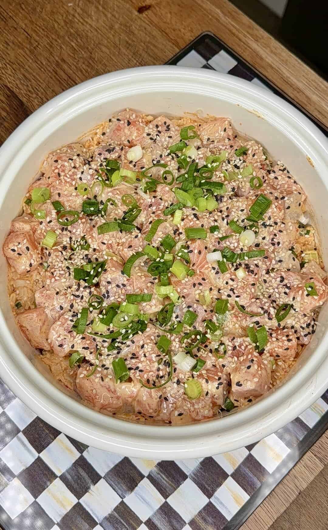 A bowl filled with diced raw fish, topped with chopped green onions, sesame seeds, and a creamy sauce, sits on a checkered mat on a wooden surface.