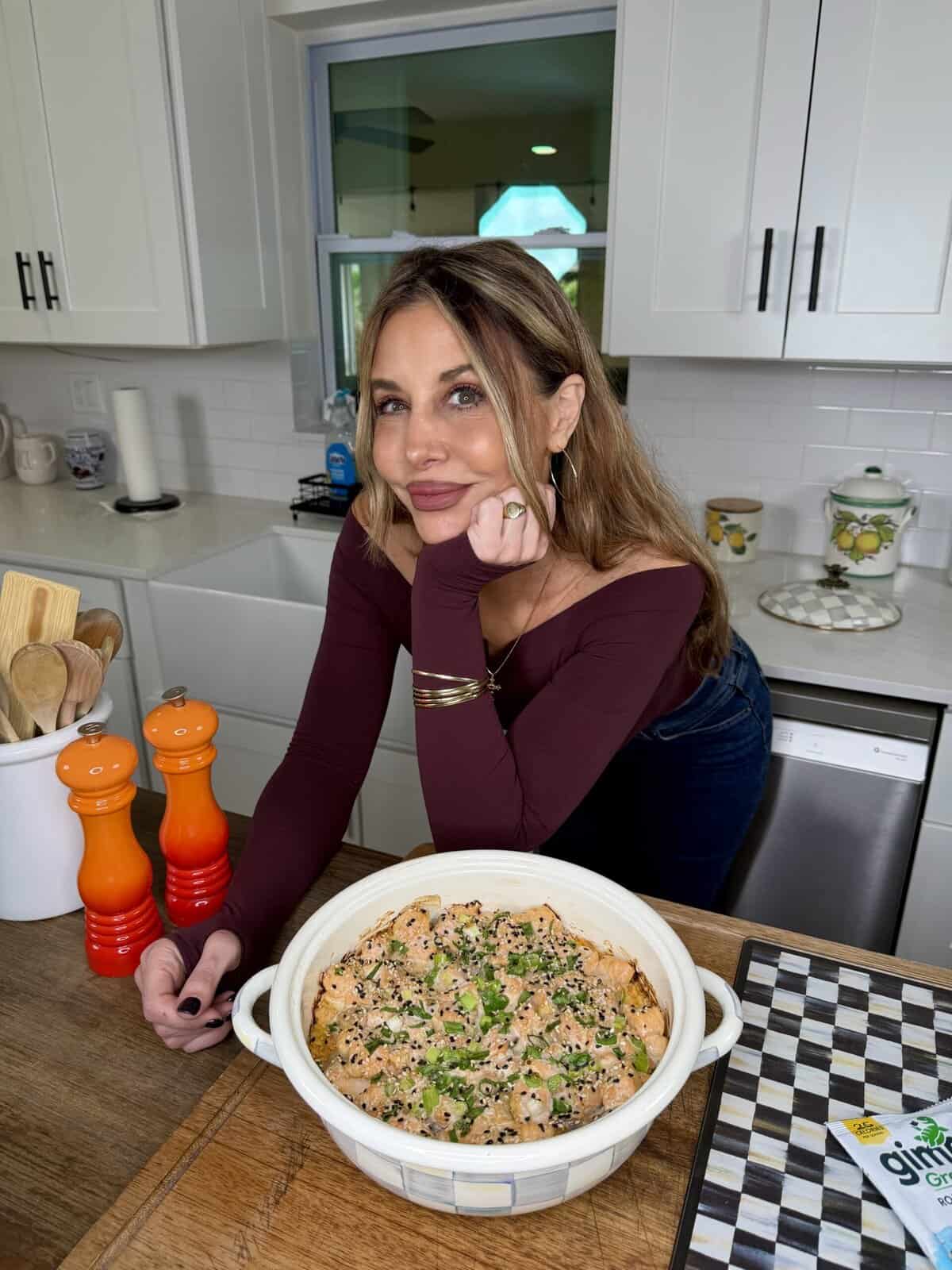 A woman with long brown hair and a burgundy top leans on a kitchen counter, smiling beside a round dish of baked food garnished with herbs. The kitchen has white cabinets, utensils, and orange pepper grinders.