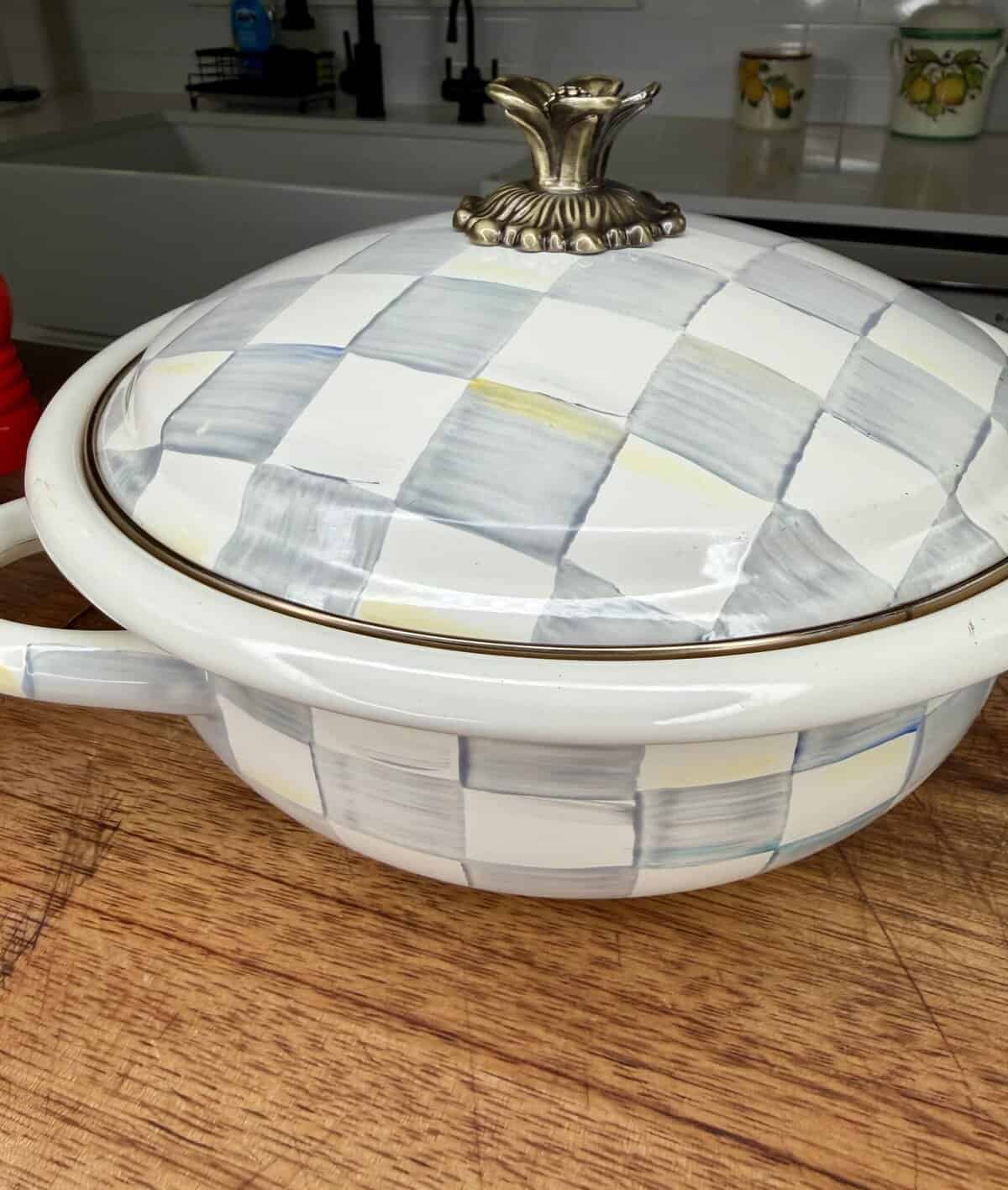 A white ceramic casserole dish with a checkered blue, gray, and yellow pattern, featuring a decorative gold handle on the lid, sits on a wooden countertop in a kitchen.