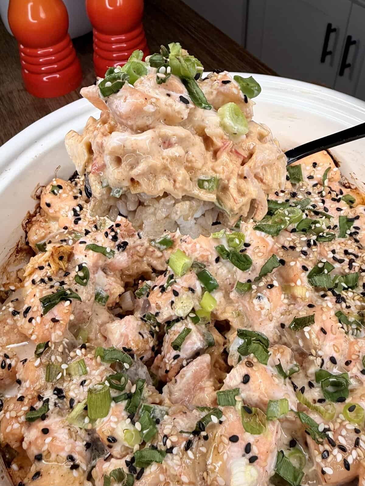 A creamy baked sushi casserole is scooped from a white dish, topped with green onions, black and white sesame seeds, and pieces of salmon. Two red containers sit in the background on a wooden counter.