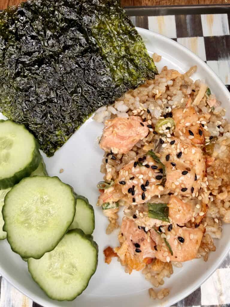 A plate with sliced cucumber, a sheet of seaweed, and a serving of rice topped with spicy salmon chunks and sprinkled with black and white sesame seeds.