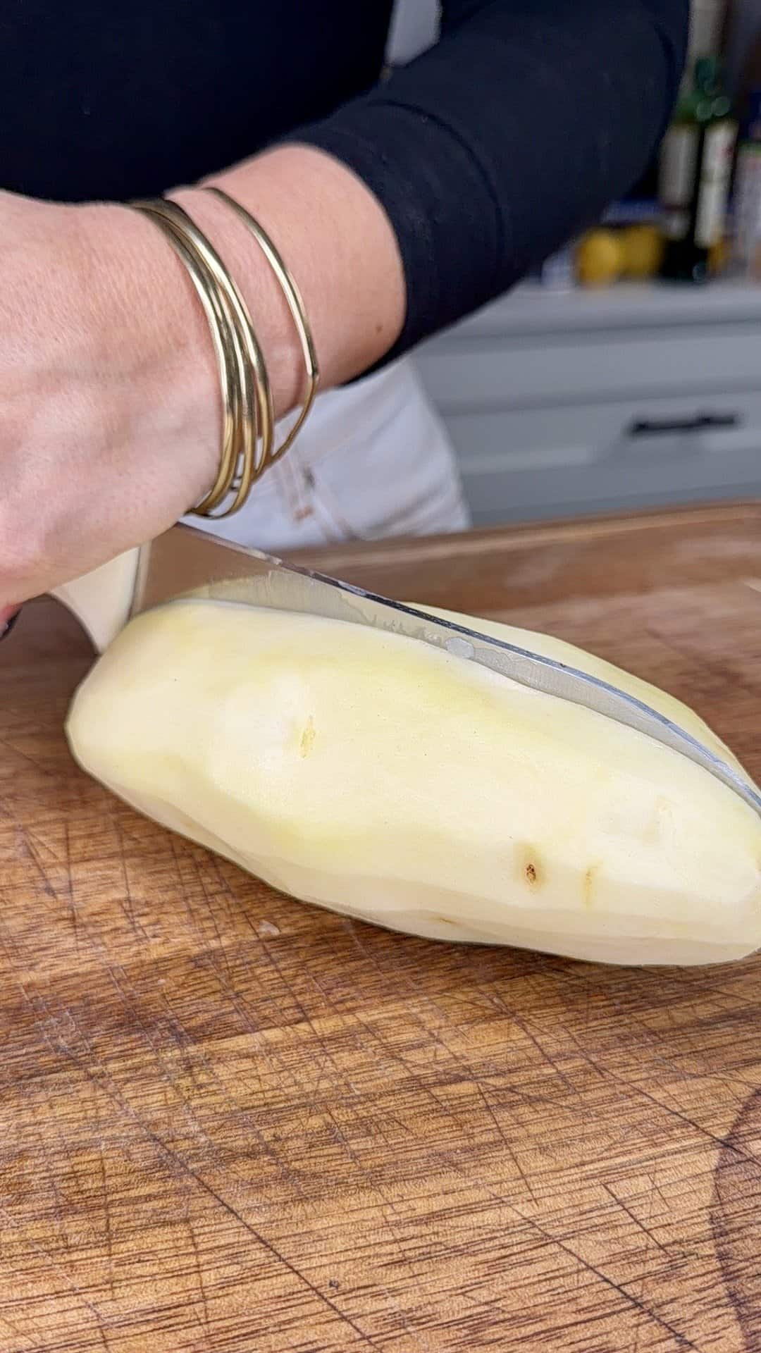 A person wearing gold bangles slices a peeled potato on a wooden cutting board, preparing it in a kitchen for Greek Restaurant-Style Lemon Roasted Potatoes.