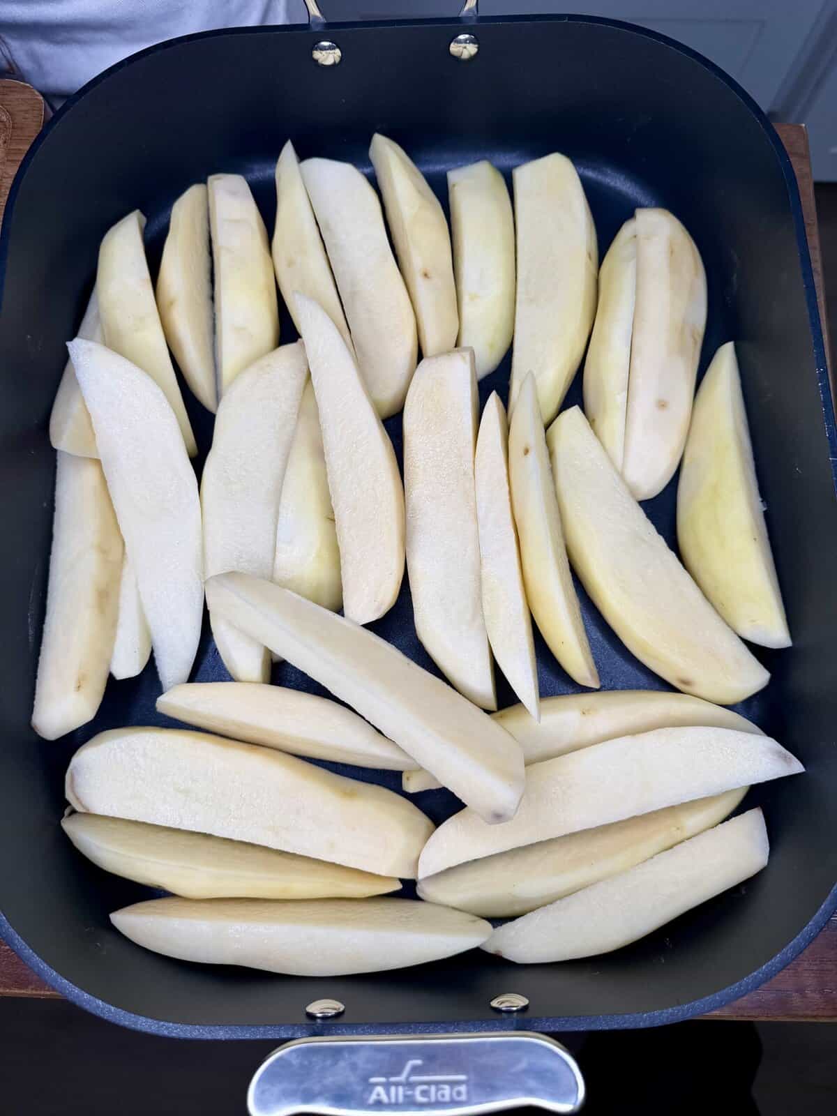 A black pan filled with evenly arranged raw potato wedges, ready for cooking. These peeled and thickly sliced potatoes await transformation into Greek Restaurant-Style Lemon Roasted Potatoes, covering the bottom of the pan.