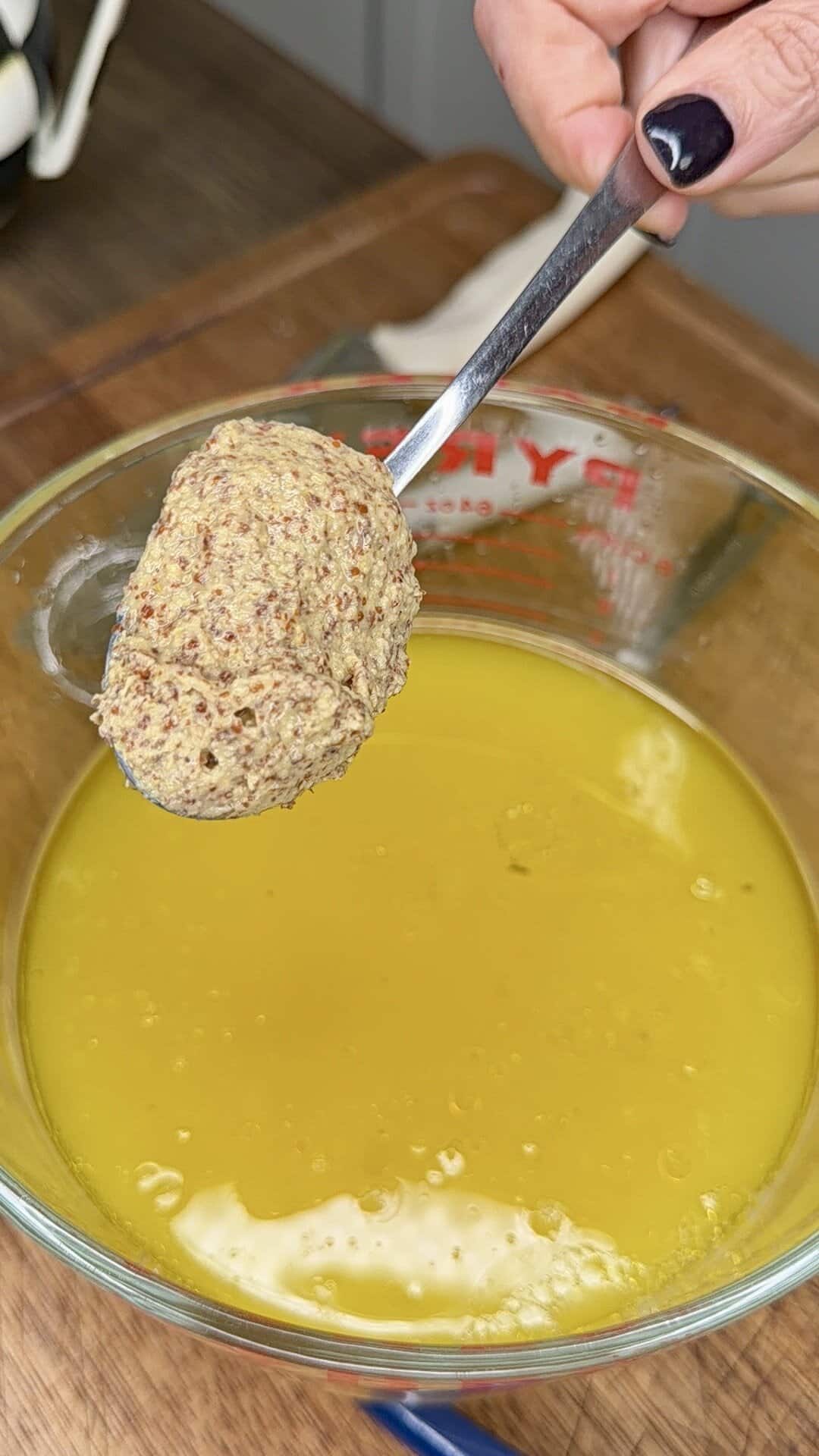 A hand holding a spoonful of coarse mustard above a bowl of yellow liquid—possibly melted butter or oil—on a wooden surface, reminiscent of preparing Greek Restaurant-Style Lemon Roasted Potatoes.