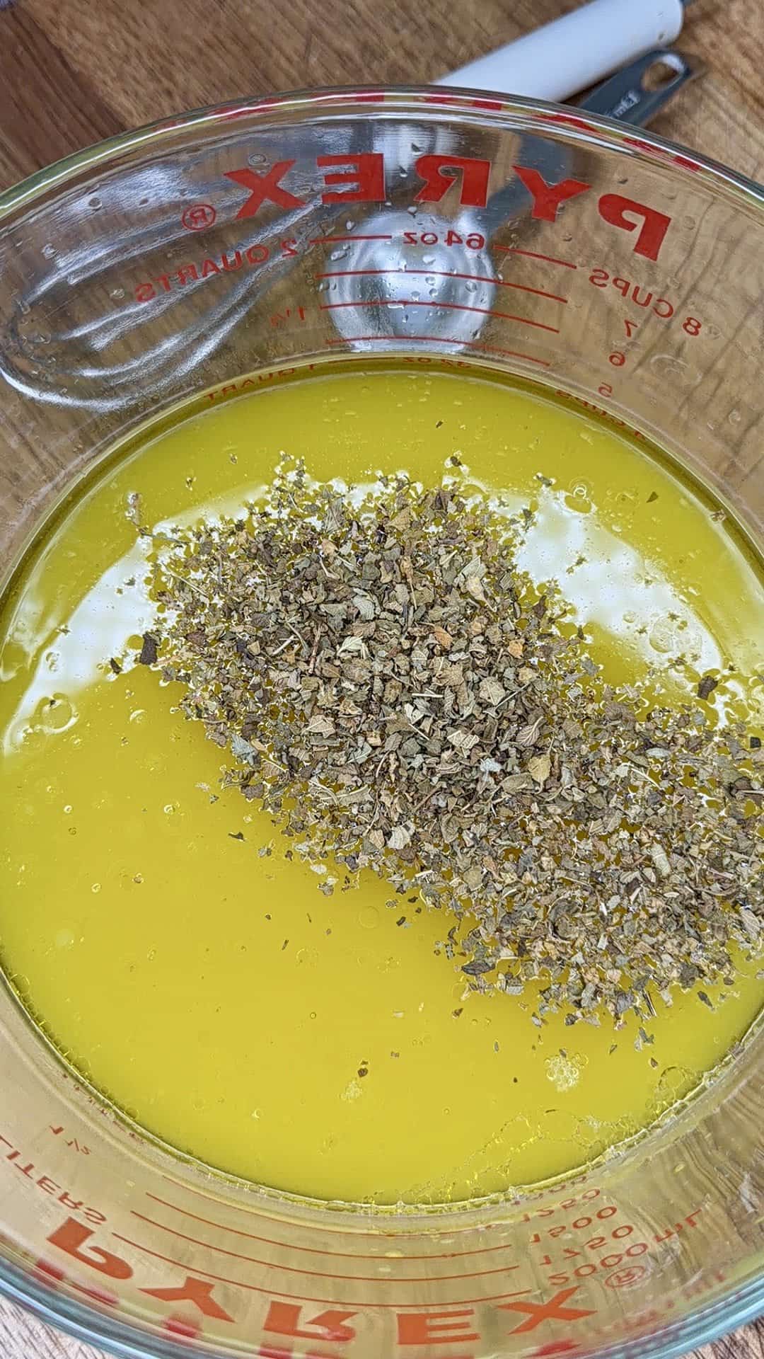 A glass Pyrex measuring cup filled with melted yellow butter and topped with dried herbs, perfect for making Greek Restaurant-Style Lemon Roasted Potatoes, sits on a wooden surface with a metal spoon in the background.