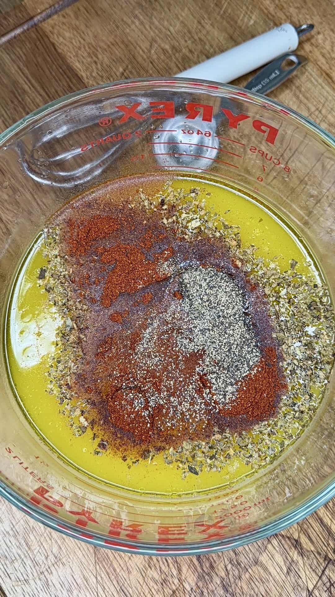 A glass measuring cup filled with yellow liquid, topped with piles of dried herbs and spices like black pepper and paprika, sits on a wooden surface—just the start for Greek Restaurant-Style Lemon Roasted Potatoes. A peeler is partially visible in the background.