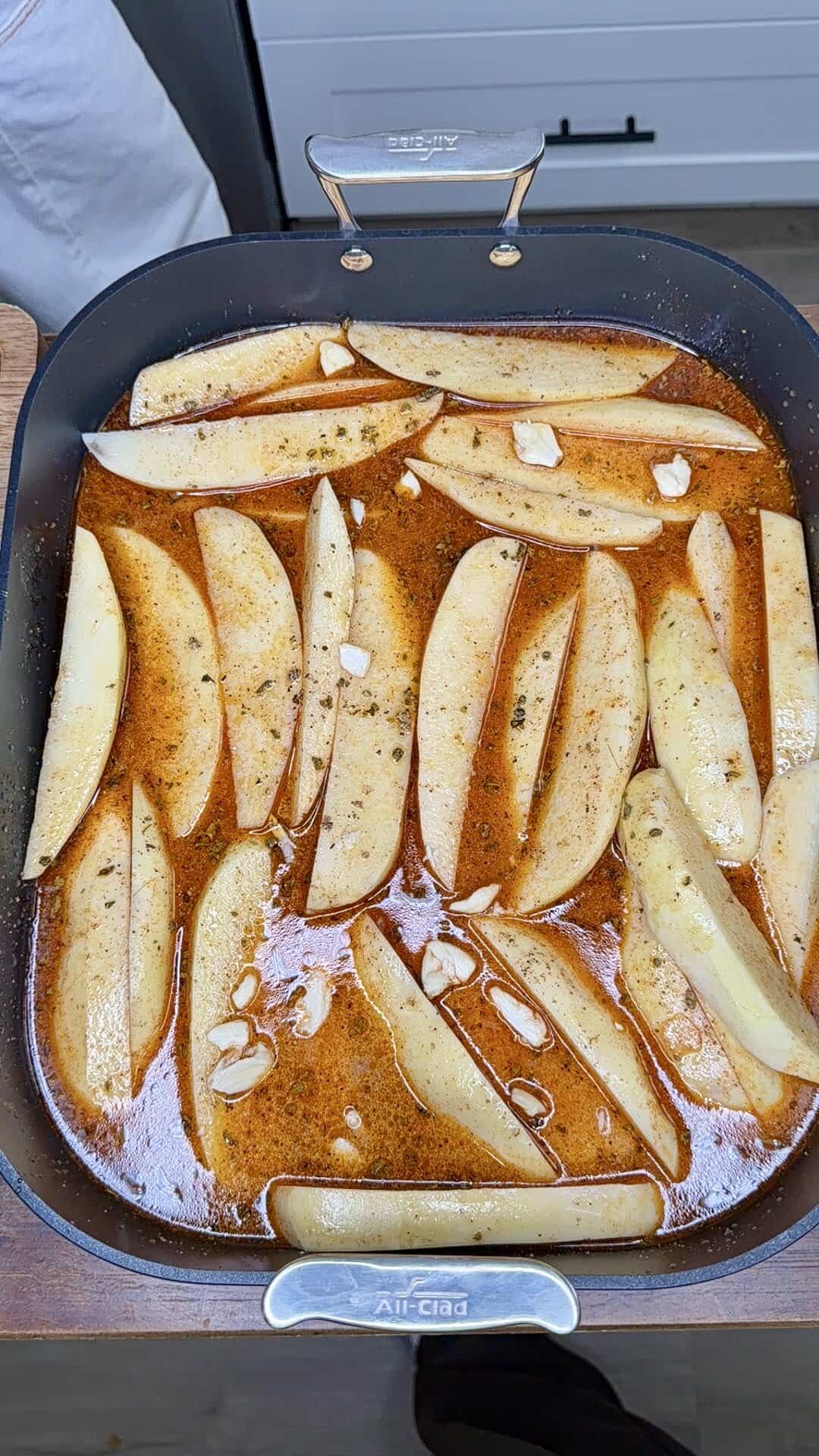 Sliced potatoes are arranged in a large All-Clad pan, partially submerged in a seasoned broth with visible herbs and garlic slices, capturing the essence of Greek Restaurant-Style Lemon Roasted Potatoes. The pan sits on a wooden surface in a bright kitchen.