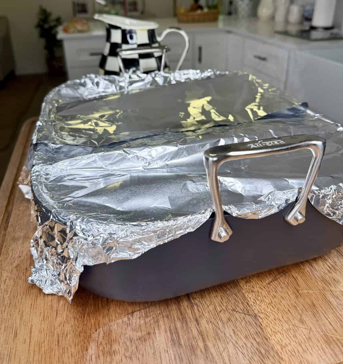 A large black roasting pan covered with aluminum foil sits on a wooden surface in a bright kitchen, ready for Greek Restaurant-Style Lemon Roasted Potatoes. A checkered teapot and other kitchen items are visible in the background.