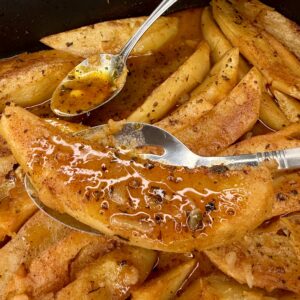 Close-up of golden Greek Restaurant-Style Lemon Roasted Potatoes in a pan, topped with herbs and sauce. A hand lifts one wedge with a serving spoon as another spoon drizzles tangy sauce over the potatoes.