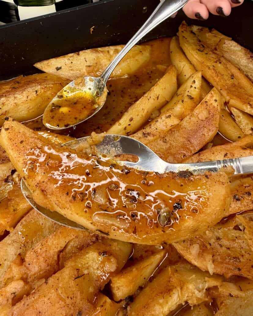 Close-up of golden Greek Restaurant-Style Lemon Roasted Potatoes in a pan, topped with herbs and sauce. A hand lifts one wedge with a serving spoon as another spoon drizzles tangy sauce over the potatoes.