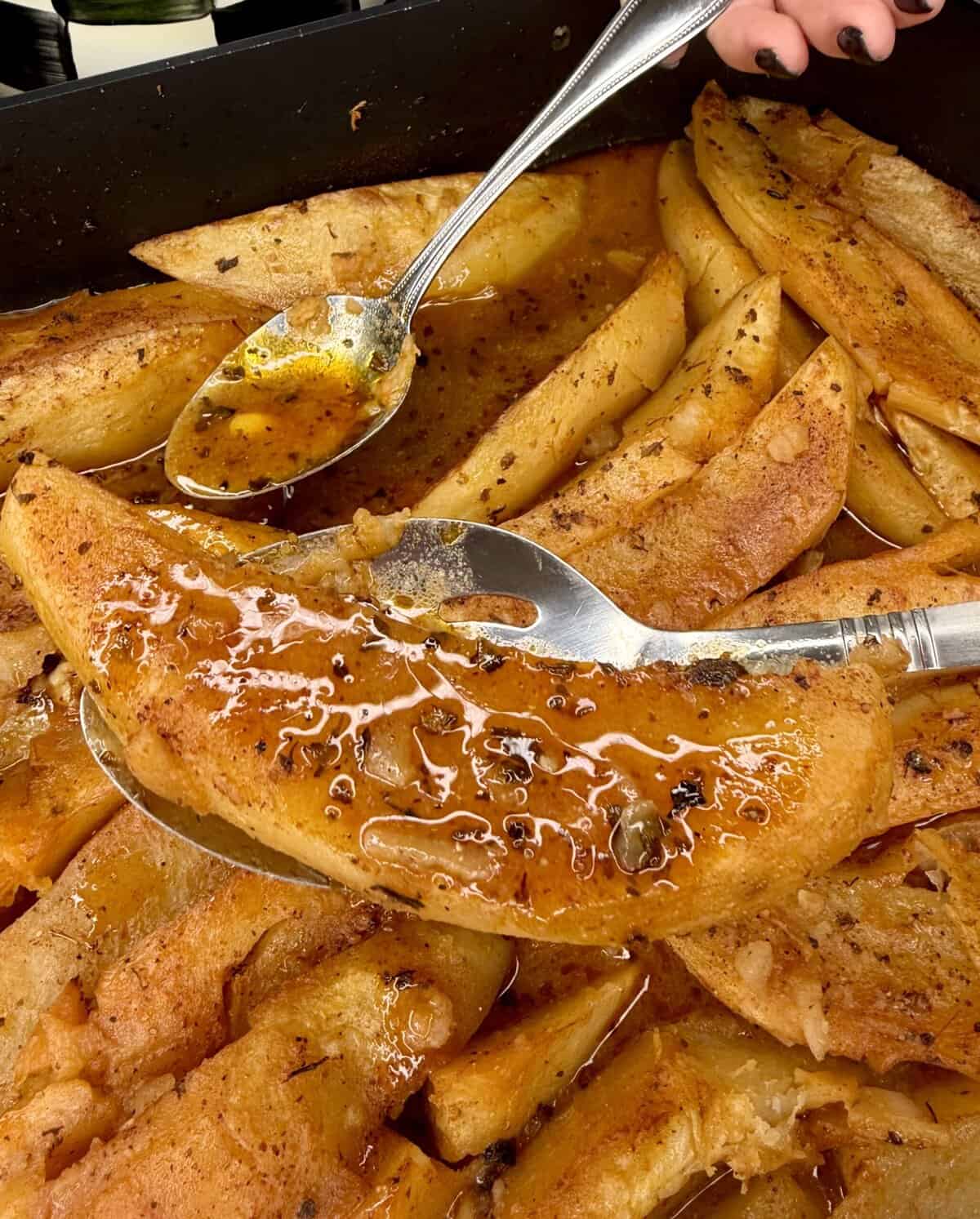 Close-up of a tray filled with golden, Greek Restaurant-Style Lemon Roasted Potatoes. A hand lifts a crispy wedge with a spoon while savory sauce is drizzled over the seasoned, well-cooked potatoes for extra flavor.