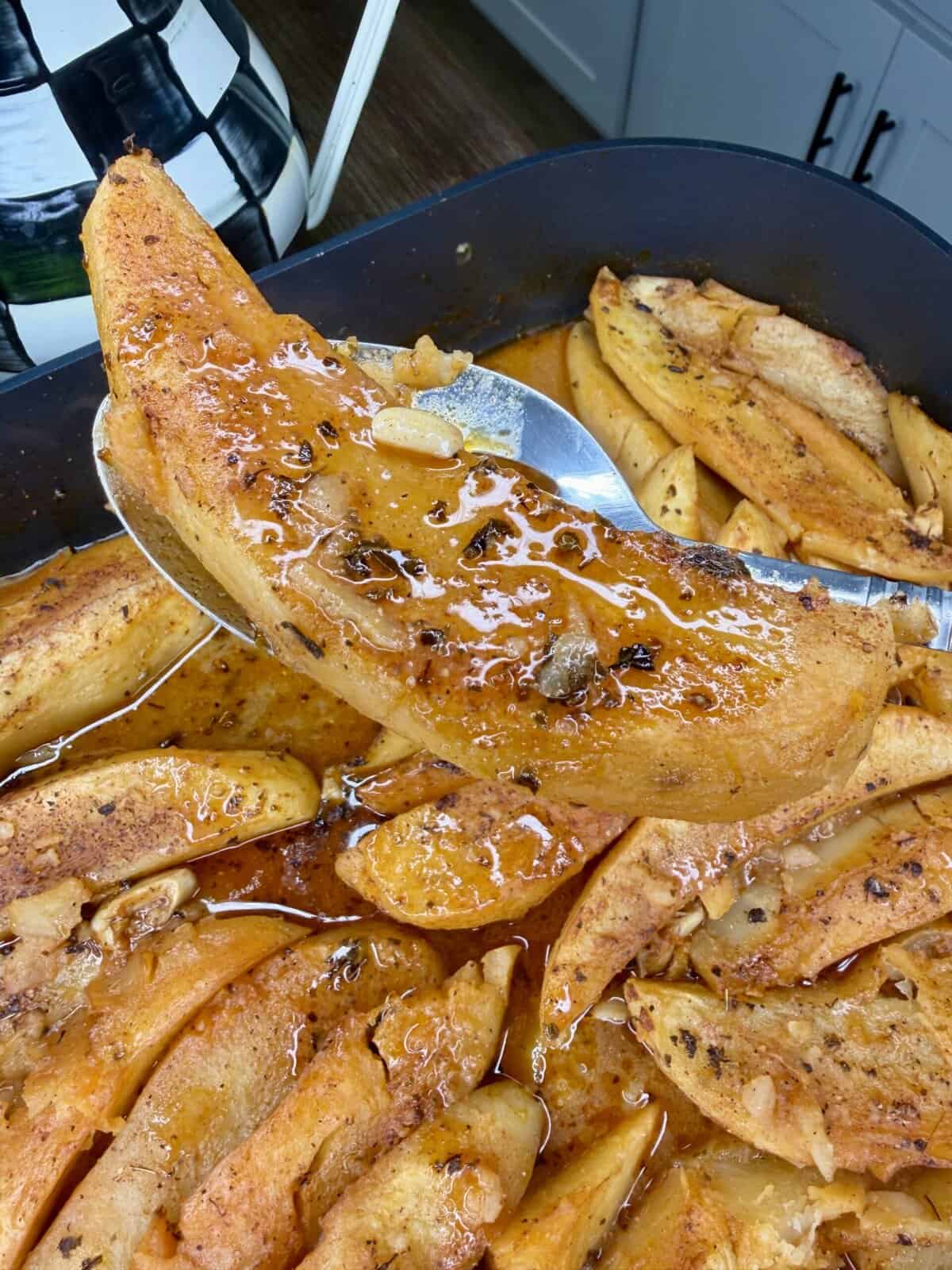 A close-up of baked sweet potato wedges in a pan, covered in a glossy, spiced glaze reminiscent of Greek Restaurant-Style Lemon Roasted Potatoes. A spoon lifts one wedge, showing its tender texture. Kitchen counters and cabinets appear in the background.