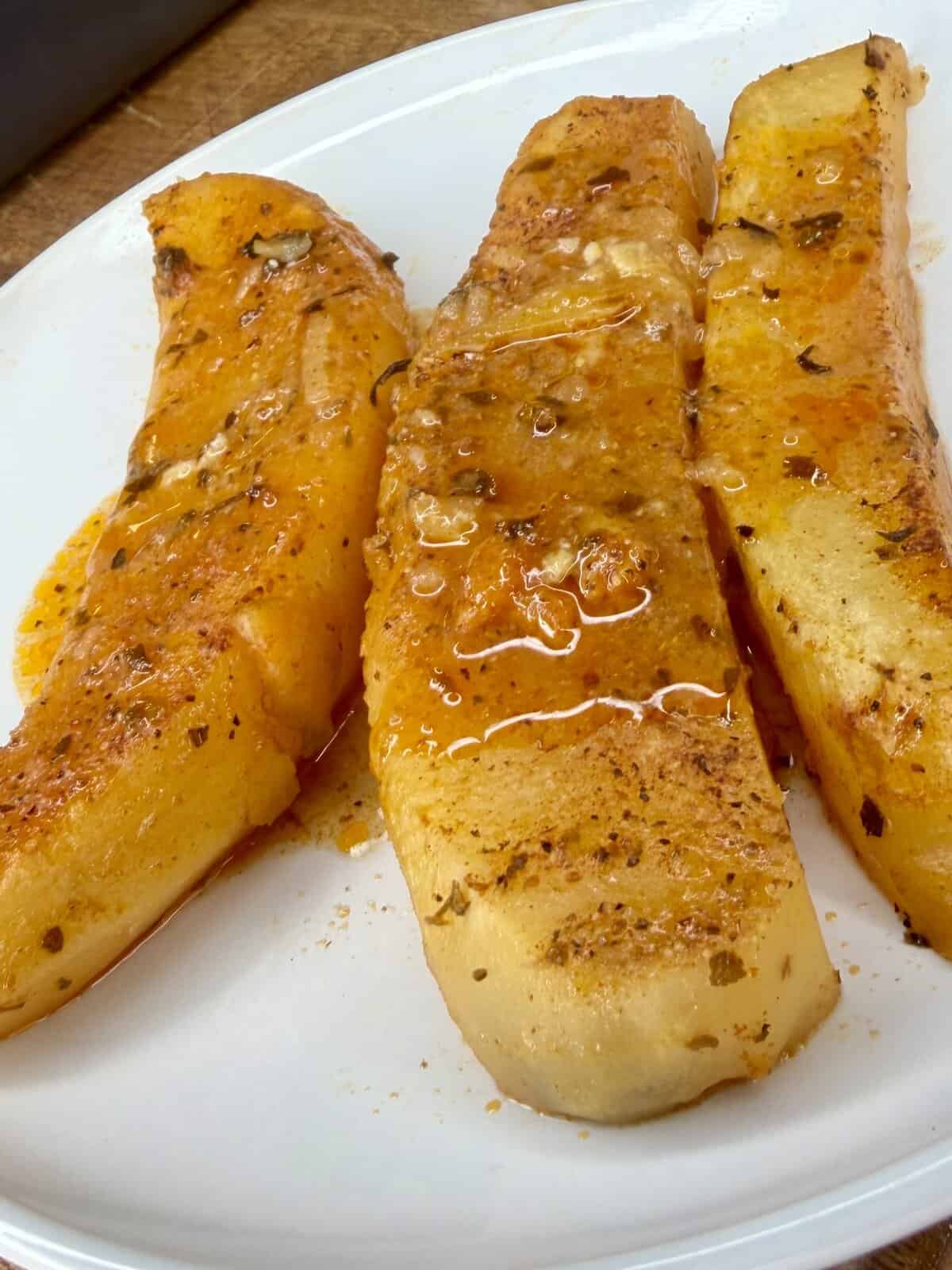 Three pieces of cooked yellow squash, inspired by Greek Restaurant-Style Lemon Roasted Potatoes, are seasoned with spices and drizzled with oil, then artfully arranged on a white plate.