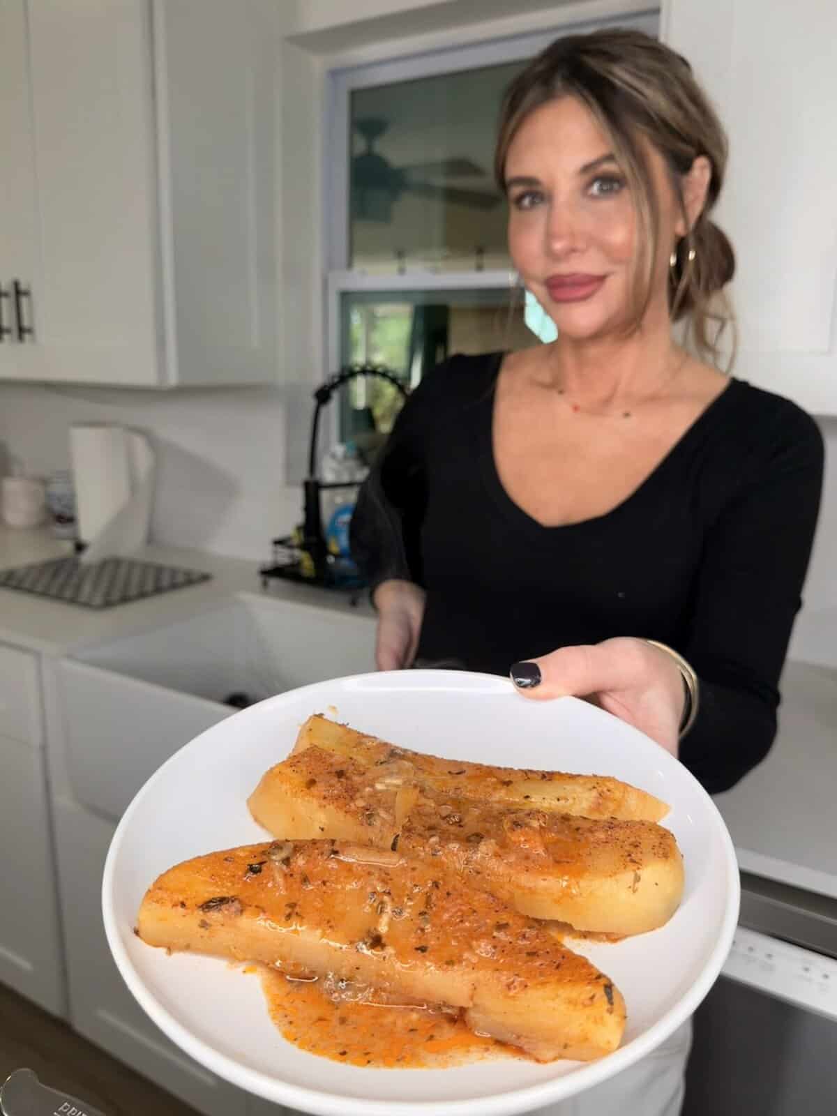 A woman in a kitchen holds a white plate with three pieces of yuca covered in a reddish sauce, garnished with herbs. She smiles and presents the dish towards the camera, just as if serving Greek Restaurant-Style Lemon Roasted Potatoes.