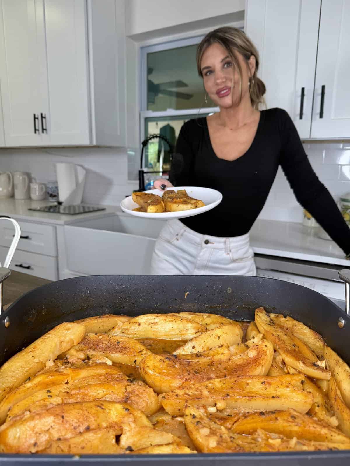A woman in a black top and white pants stands in a modern kitchen, holding a plate of Greek Restaurant-Style Lemon Roasted Potatoes. In the foreground is a large pan filled with perfectly seasoned, roasted potatoes.