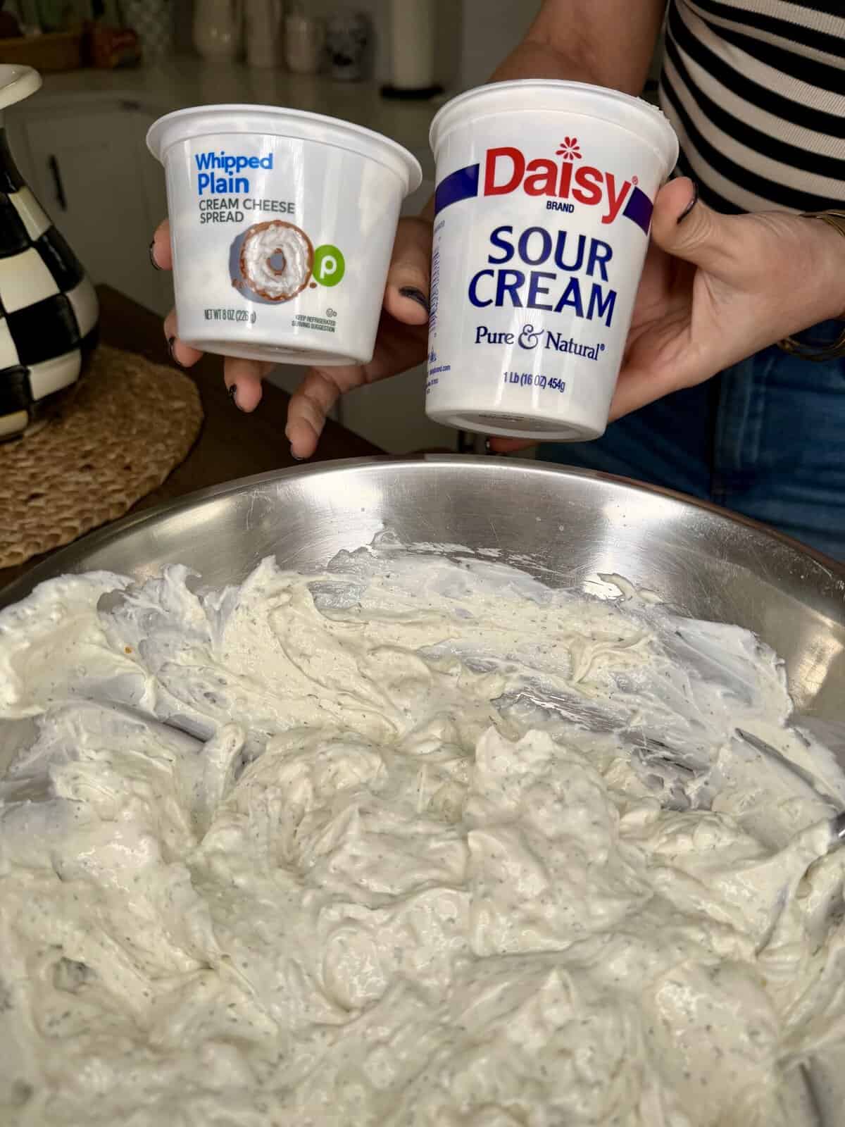 A person holds containers of Publix whipped plain cream cheese spread and Daisy sour cream above a metal bowl filled with a creamy, herb-speckled mixture, in a kitchen setting.