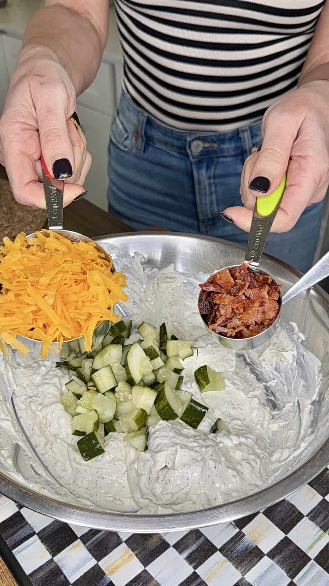 A person holds measuring cups of shredded cheddar cheese and chopped bacon over a mixing bowl filled with creamy mixture and diced pickles, preparing to add the ingredients.