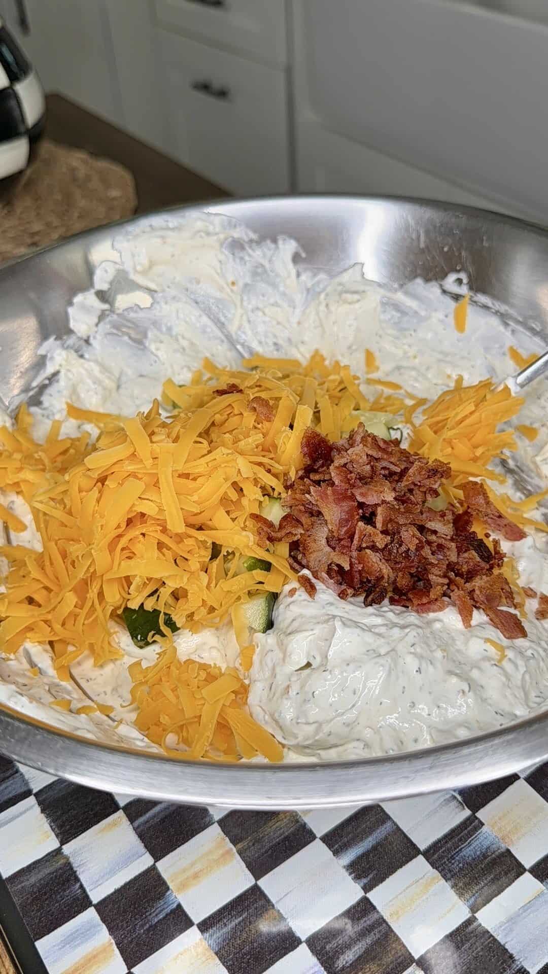 A metal mixing bowl filled with creamy dip, topped with shredded cheddar cheese, chopped bacon, and sliced green onions. A spoon is partially visible in the mixture, and the bowl sits on a checkered surface.