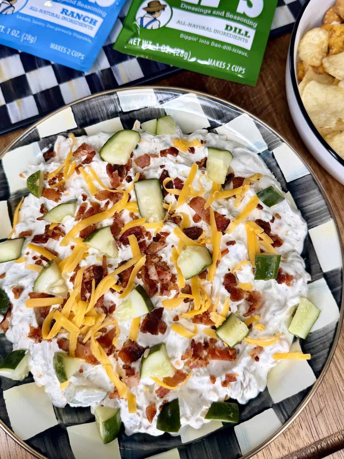 A bowl of creamy dip topped with chopped bacon, shredded cheddar cheese, and cucumber pieces, surrounded by bags of seasoning mix and a bowl of potato chips.