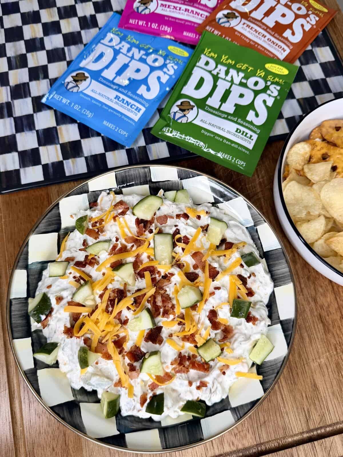 A bowl of dip topped with shredded cheese, bacon bits, and cucumber pieces sits on a table next to a bowl of potato chips and three colorful packets of Dinos Dips seasoning mixes.
