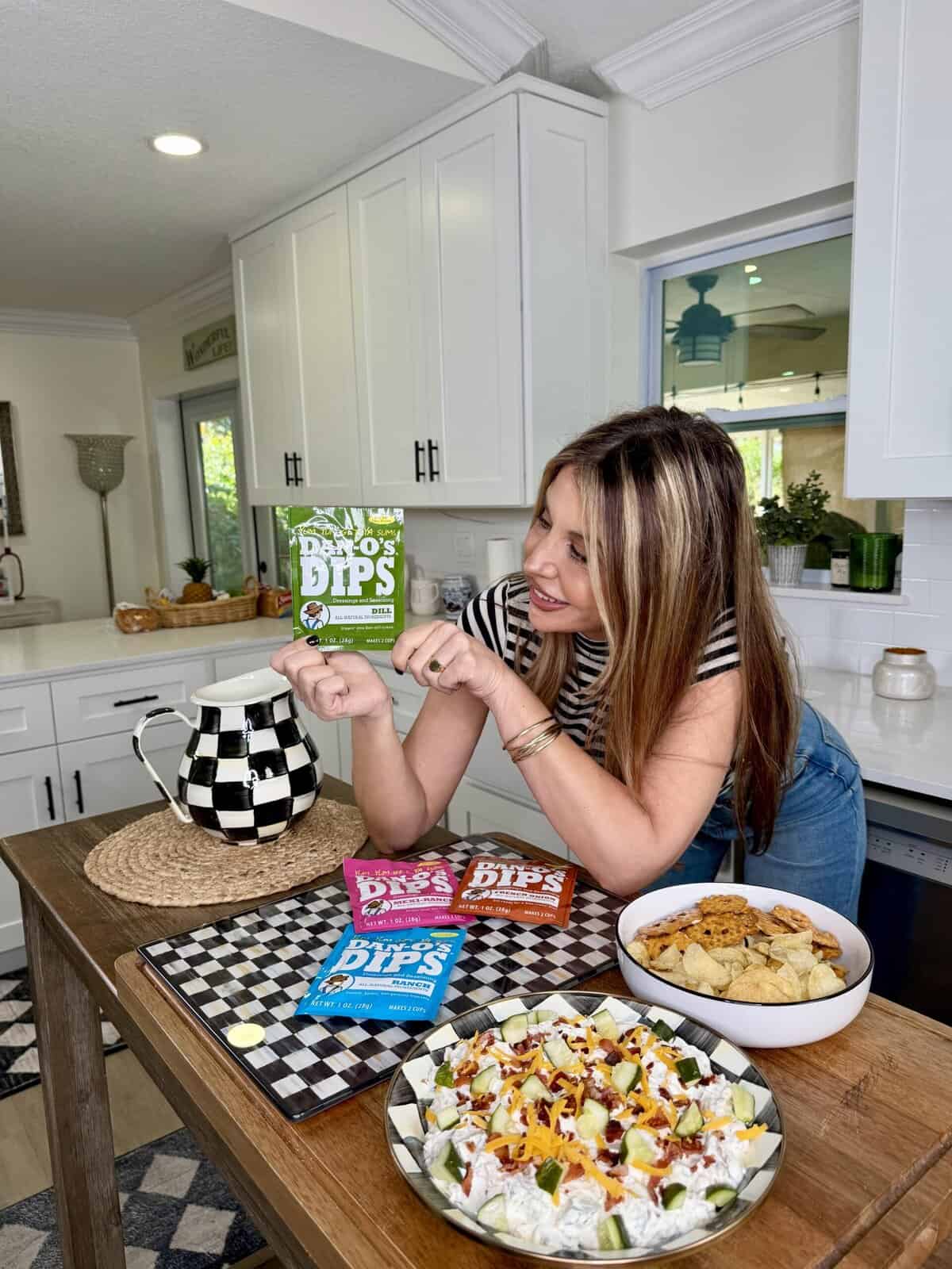 A woman in a kitchen smiles while holding a green packet of Dan-O’s Dips. Three other dip packets are on the counter next to a bowl of chips and a plate of nachos. The kitchen is bright and modern.