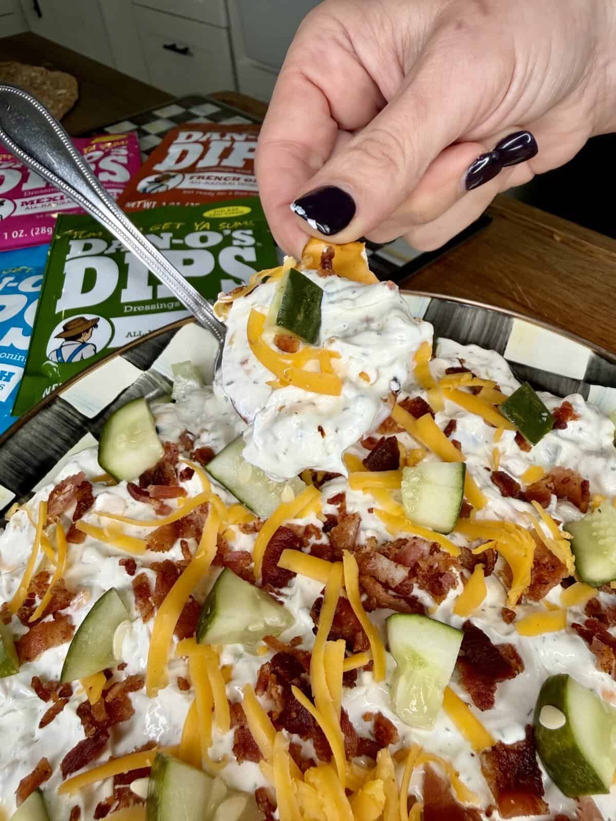 A hand with dark-painted nails scoops a cheesy bacon dip with cucumber from a plate using a spoon. The dip is topped with shredded cheese, bacon bits, and cucumber pieces. Colorful dip mix packets are in the background.