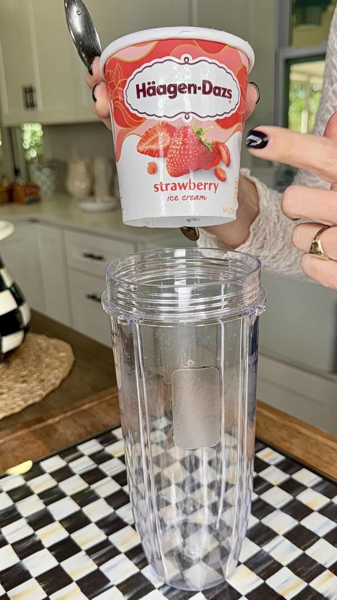 A person holds a container of Häagen-Dazs strawberry ice cream above an empty blender cup in a kitchen, inspired by Easy Strawberry Tres Leches Poke Cake, preparing to make a delicious smoothie or milkshake.