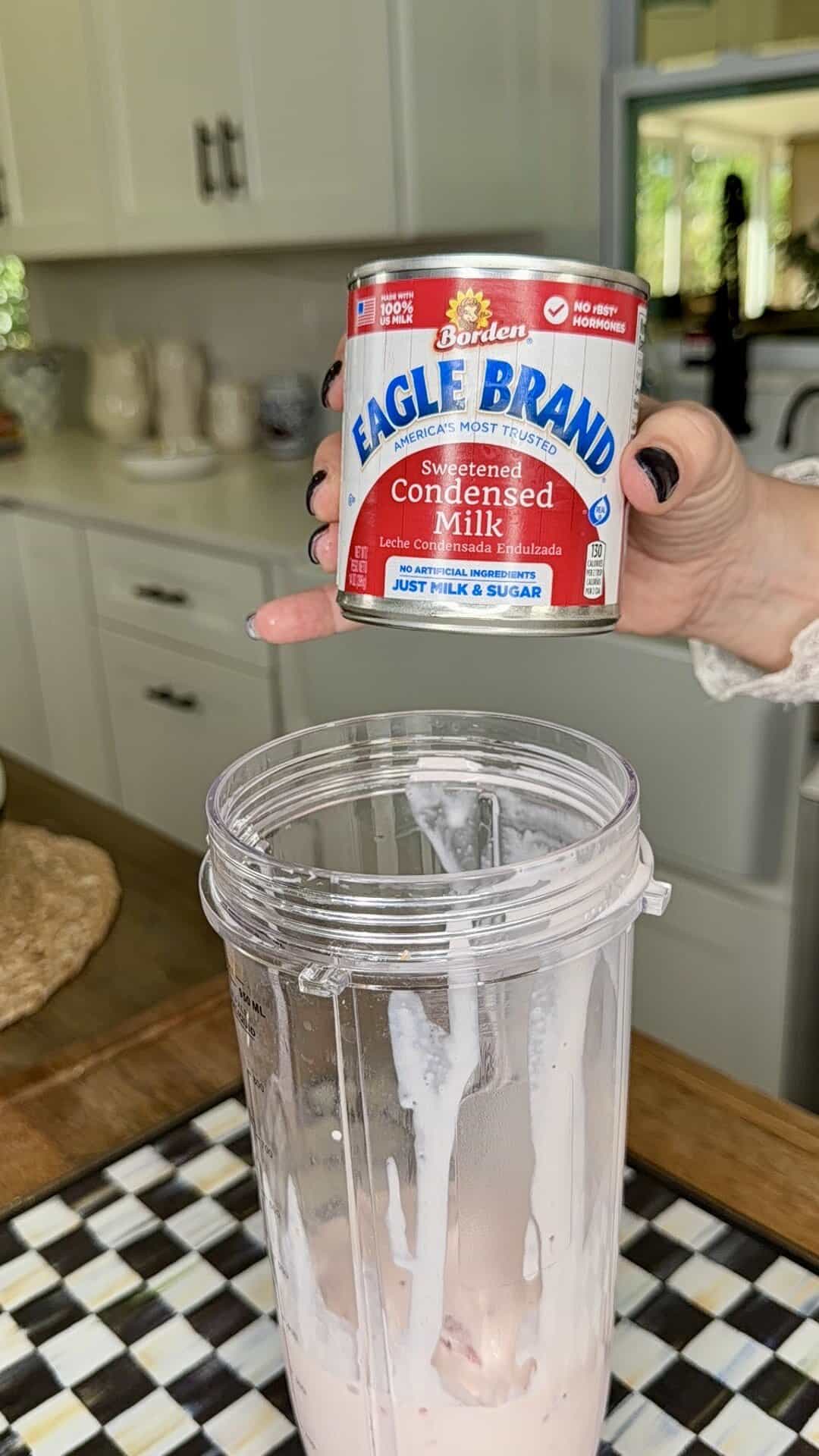 A hand holds a can of Eagle Brand sweetened condensed milk above a blender cup filled with a partially blended pink mixture, perfect for making Easy Strawberry Tres Leches Poke Cake in a kitchen setting.