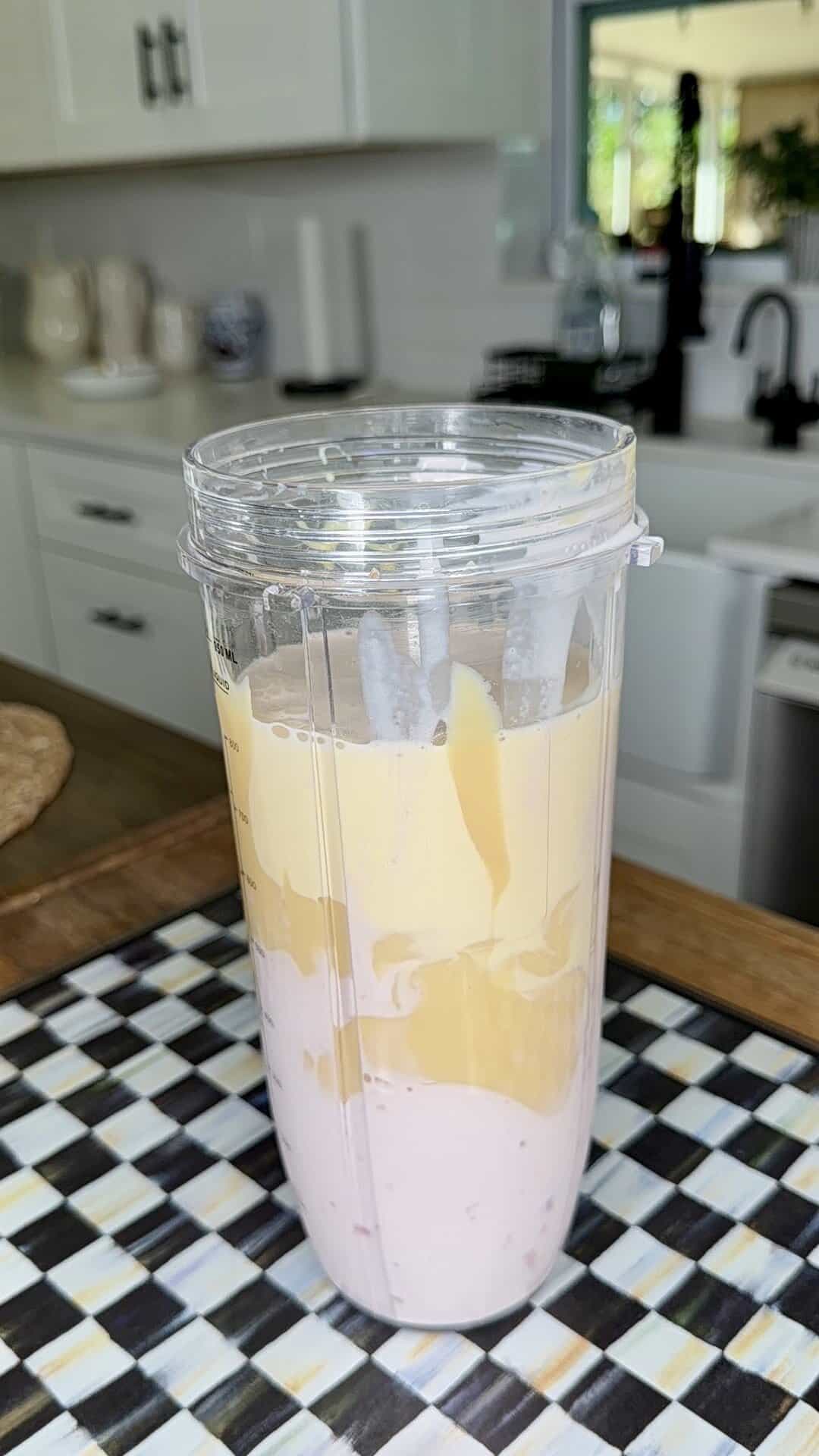 A clear blender cup filled with pink smoothie and yellow liquid sits on a black, white, and yellow checkered surface—perfect inspiration for enjoying an Easy Strawberry Tres Leches Poke Cake in a bright kitchen with white cabinets and a sink in the background.