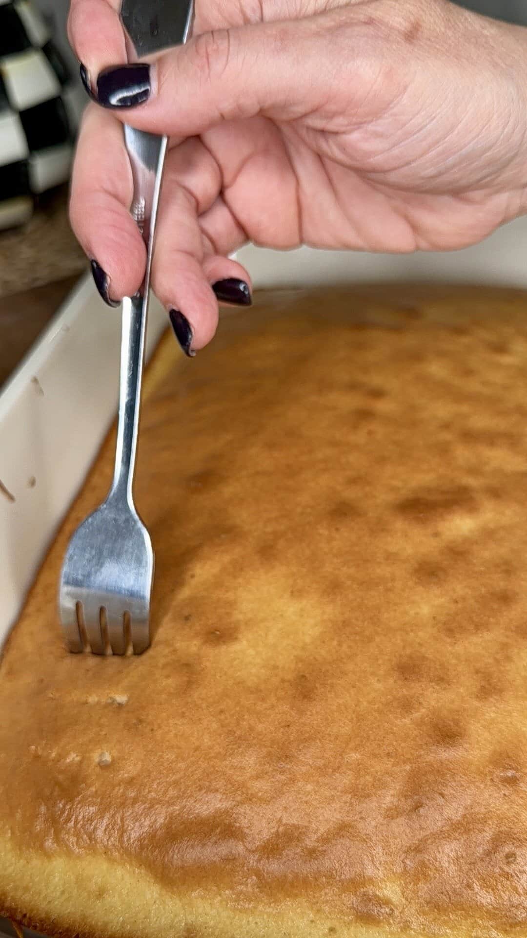 A hand with dark nail polish uses a fork to poke holes in a golden-brown Easy Strawberry Tres Leches Poke Cake in a baking pan.