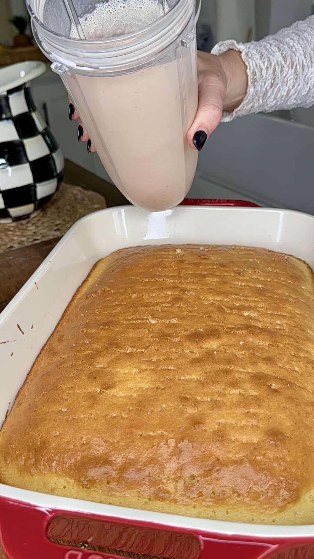 A hand holds a blender cup with a creamy liquid, pouring it over an Easy Strawberry Tres Leches Poke Cake in a red baking dish. A black and white checkered pitcher is visible in the background.