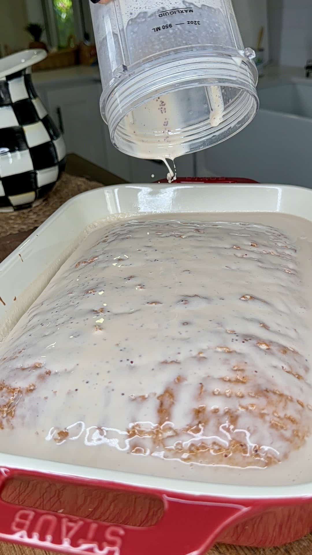 A creamy white glaze is being poured from a blender onto an Easy Strawberry Tres Leches Poke Cake in a red baking dish, with a black and white checkered teapot in the background.