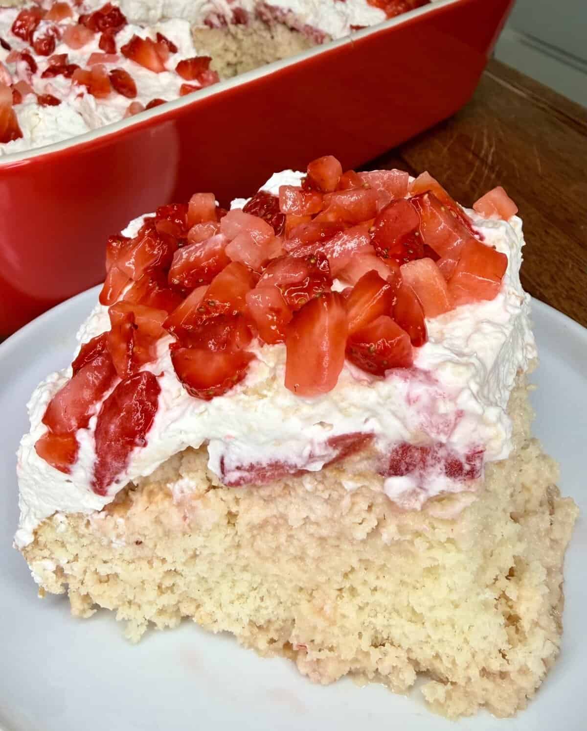 A slice of Easy Strawberry Tres Leches Poke Cake topped with whipped cream and chopped fresh strawberries sits on a white plate, with the rest of the cake visible in a red baking dish in the background.