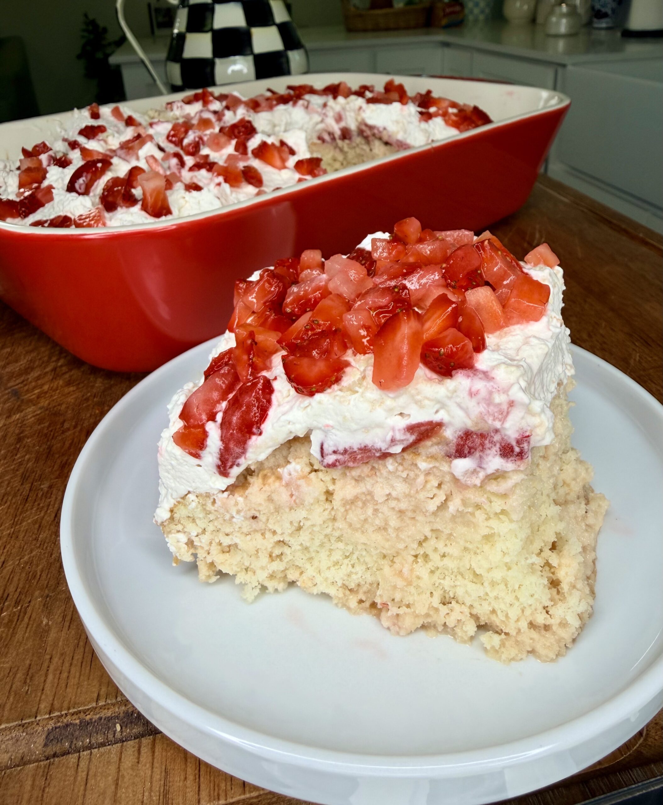 A slice of Easy Strawberry Tres Leches Poke Cake with whipped cream and chopped strawberries sits on a white plate, with the rest of the dessert in a red baking dish in the background on a wooden table.