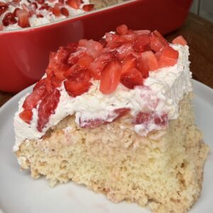 A slice of Easy Strawberry Tres Leches Poke Cake topped with whipped cream and chopped fresh strawberries sits on a white plate, with a large red dish of the same dessert in the background.