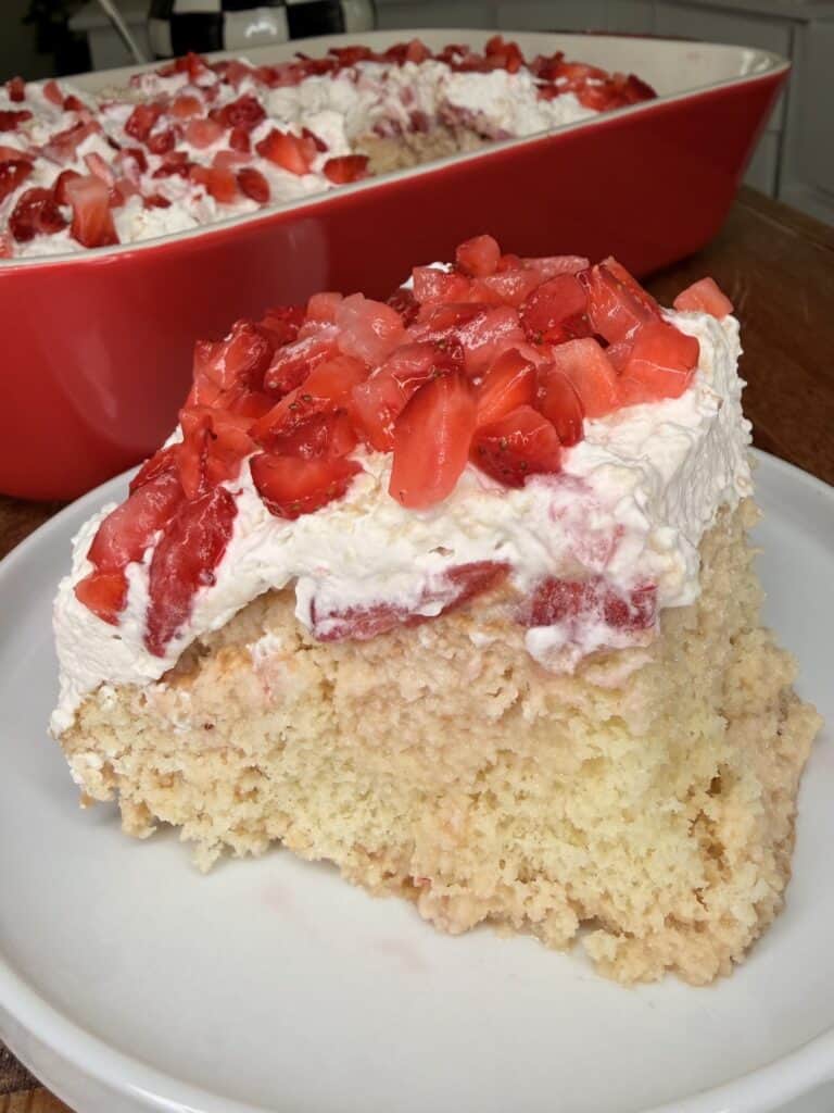 A slice of Easy Strawberry Tres Leches Poke Cake topped with whipped cream and chopped fresh strawberries sits on a white plate, with a large red dish of the same dessert in the background.