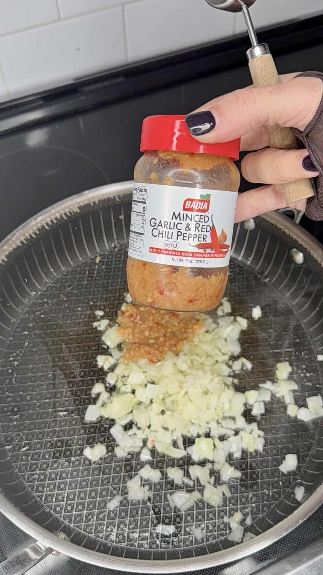 A hand holds a jar of Badia Minced Garlic & Red Chili Pepper over a pan with chopped onions sautéing on a stovetop.