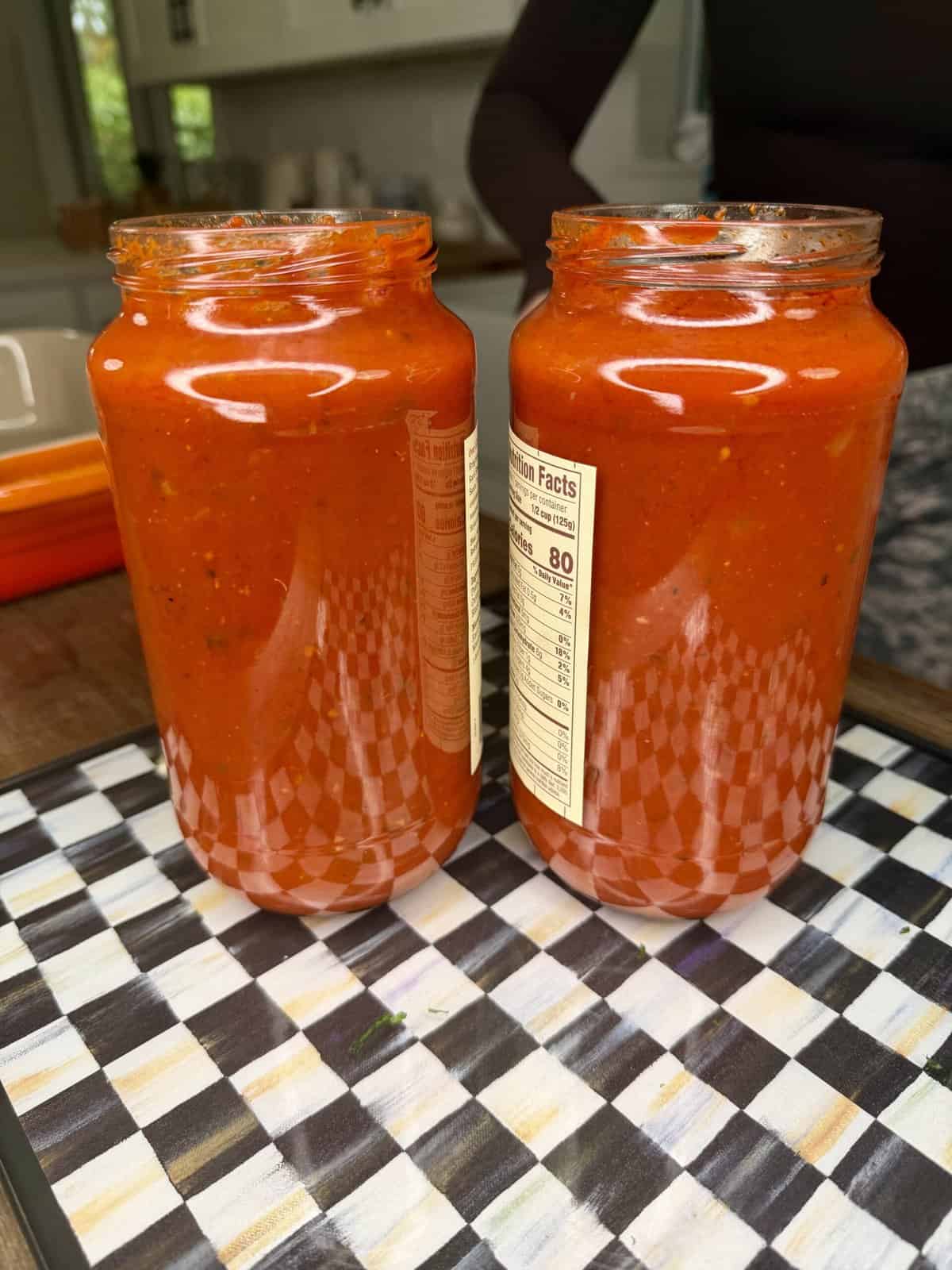 Two glass jars filled with vibrant red tomato sauce are placed side by side on a checkered tray in a kitchen setting. The jars have labels with nutritional information visible.