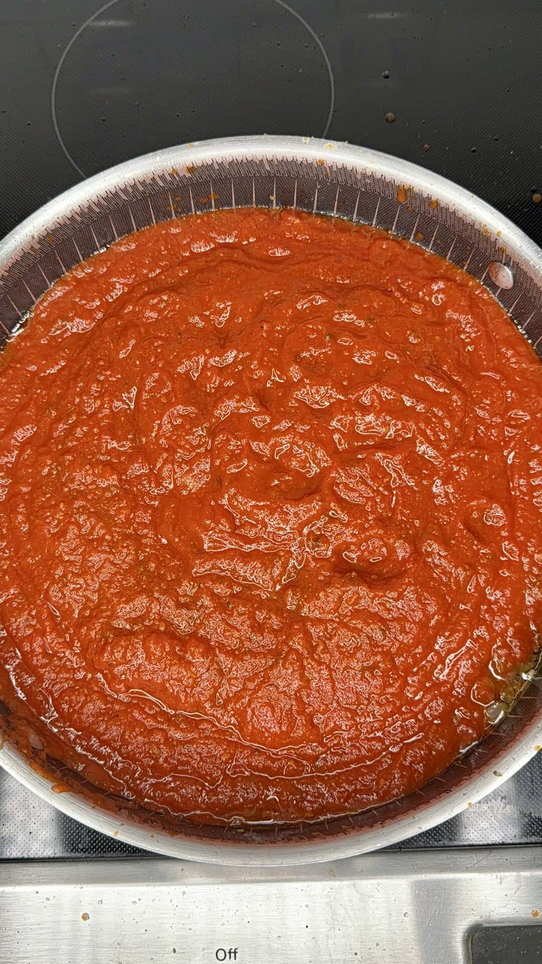 A large silver pan filled with red tomato sauce sits on a stovetop, with some green herbs visible around the edges of the sauce.
