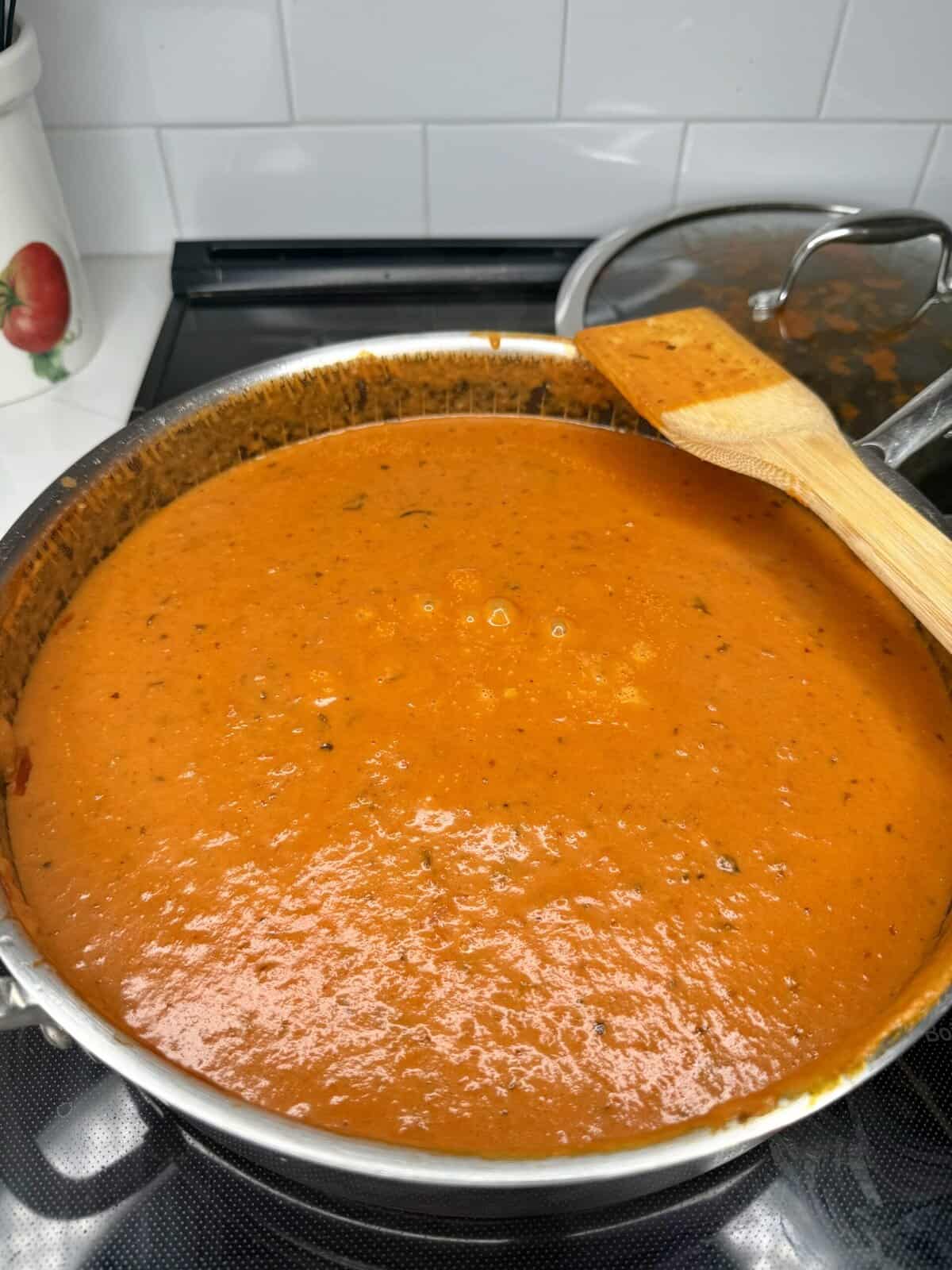 A large metal pot filled with creamy tomato soup sits on a stovetop. A wooden spatula rests across the side of the pot. A glass-lidded pot and kitchen decor are visible in the background.