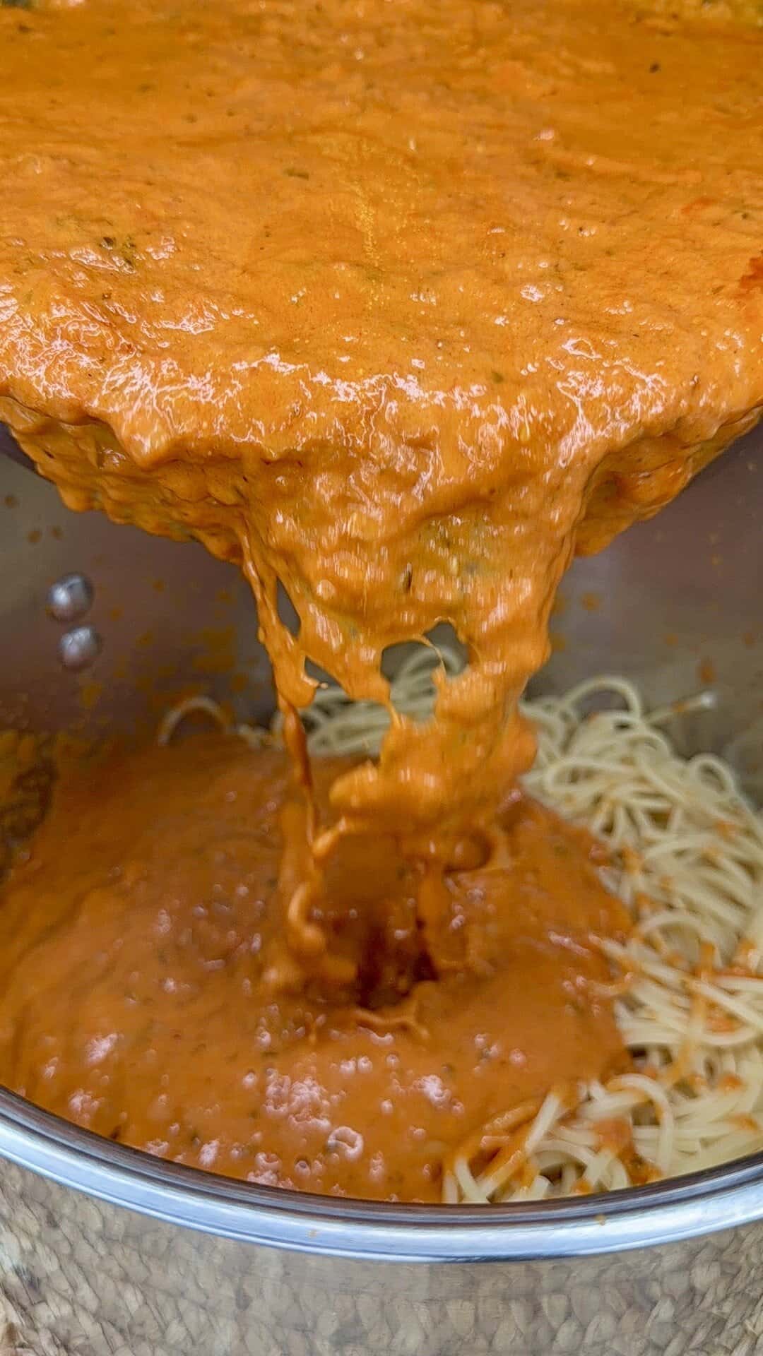 A close-up view of rich, red tomato sauce being poured over a pot of cooked spaghetti noodles. The sauce is thick and creamy, covering the pasta as it flows from the pot above.