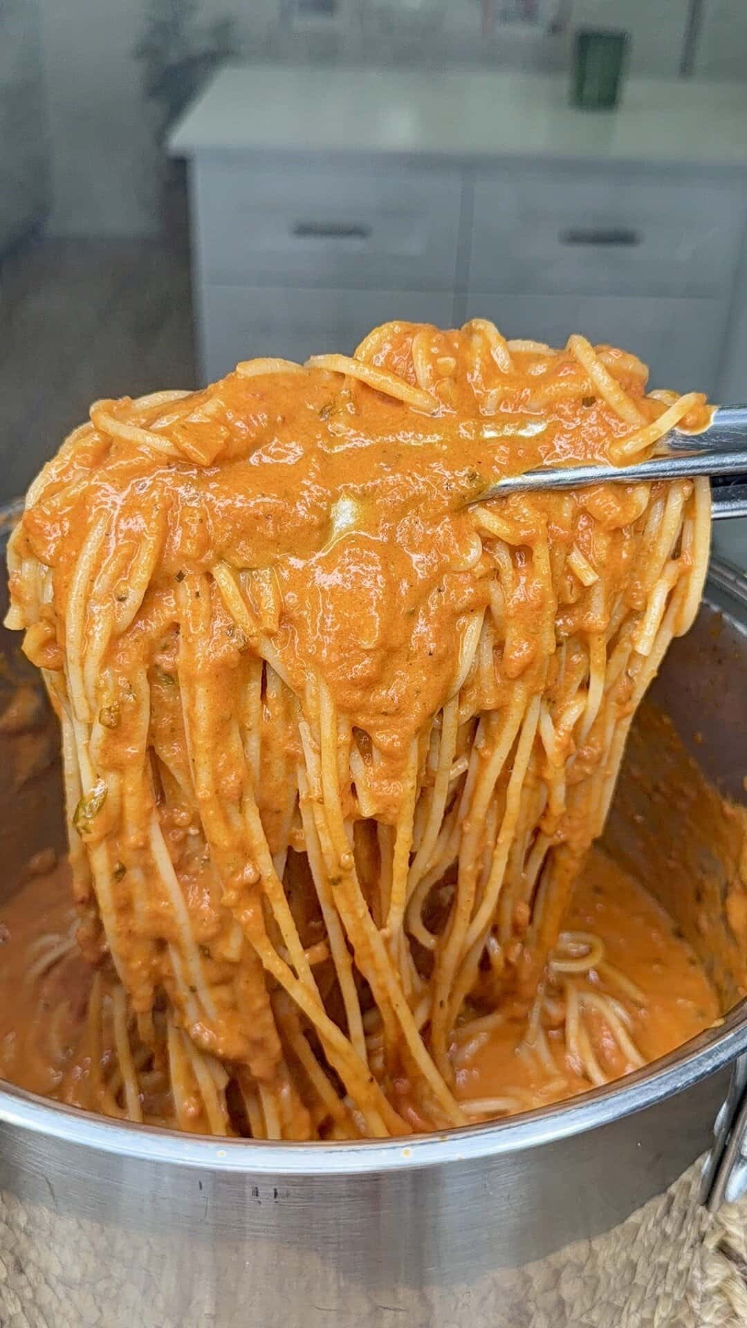 A close-up of spaghetti being lifted from a pot with tongs, coated in a thick, creamy tomato sauce. A white kitchen is slightly visible in the blurry background.