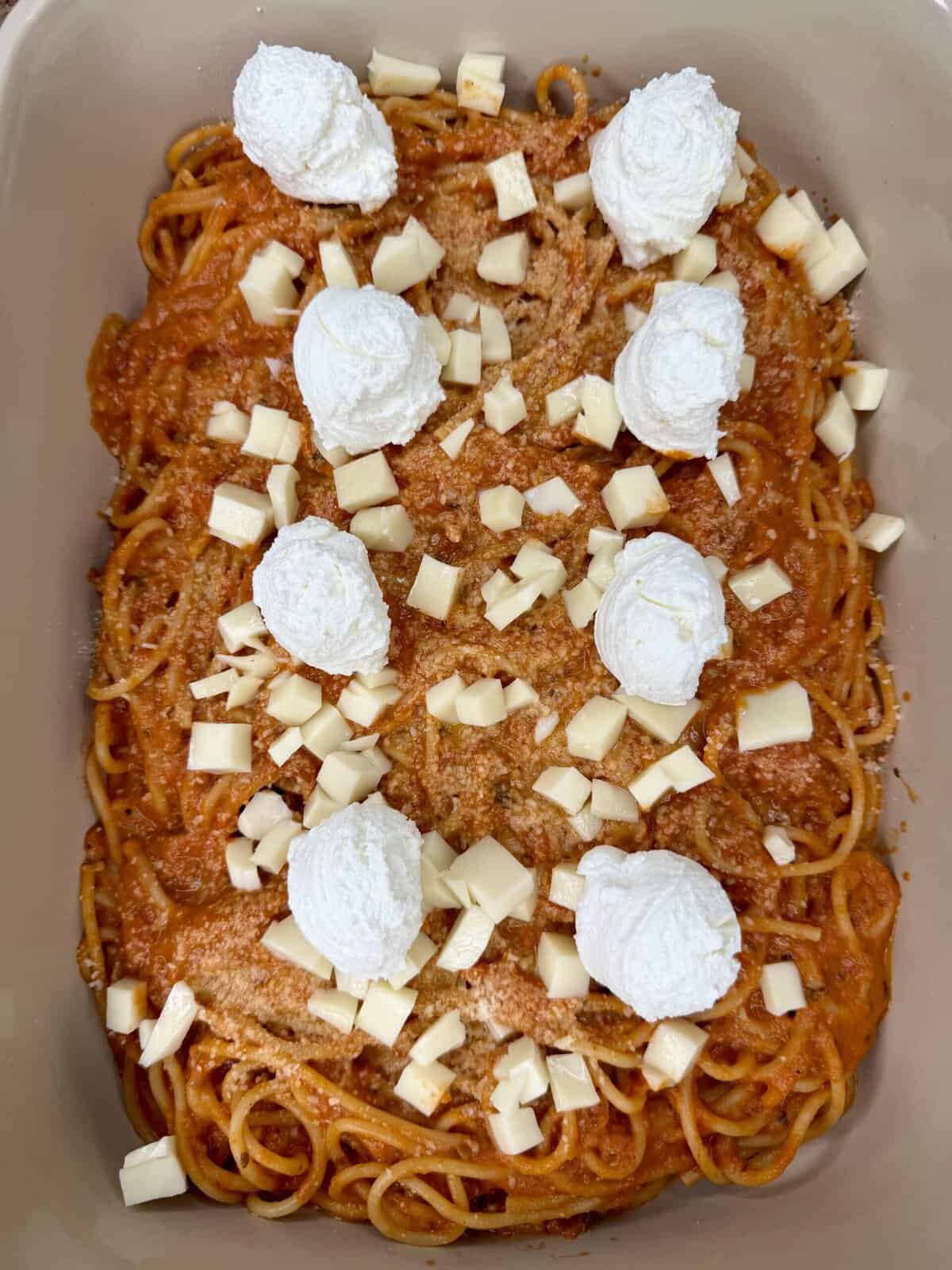 A baking dish filled with spaghetti in tomato sauce, topped with dollops of ricotta cheese and scattered cubes of mozzarella cheese.