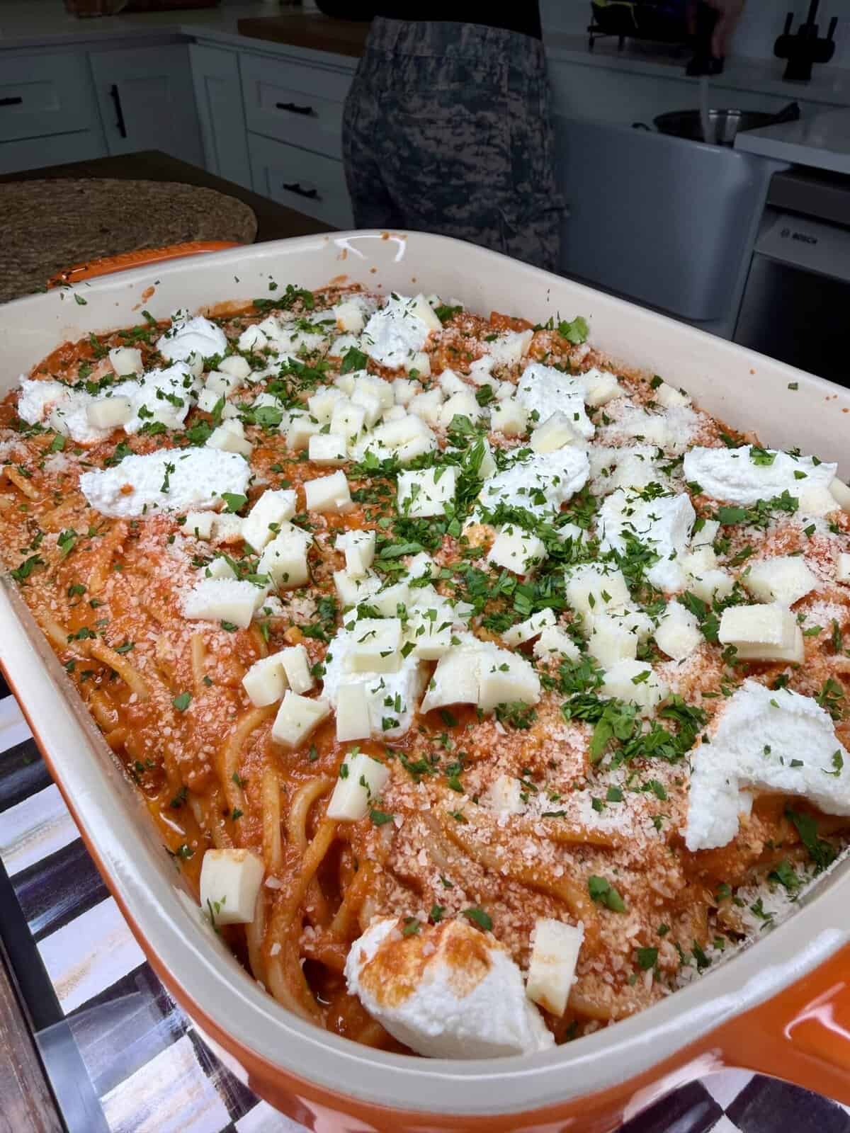 A large orange baking dish filled with spaghetti in red sauce, topped with chunks of white cheese, ricotta, and chopped herbs, sits on a kitchen counter. A person stands in the background near a stove and sink.