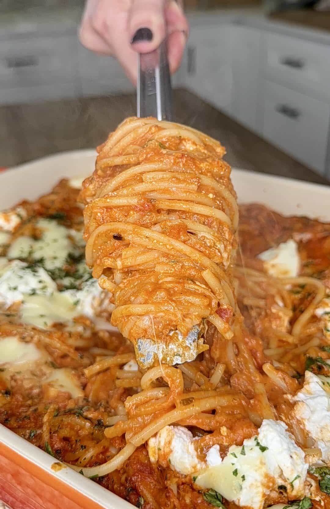 A hand holding tongs lifts a serving of cheesy baked spaghetti with tomato sauce from a casserole dish, with steam rising and melted cheese visible throughout.
