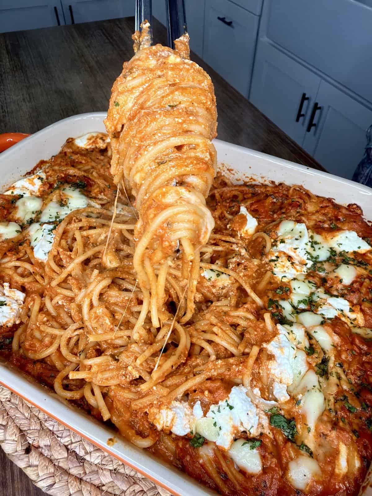 A close-up of spaghetti baked in tomato sauce with melted cheese and herbs in a casserole dish. A serving of spaghetti is being lifted with tongs, showing stretchy cheese. White cabinets are visible in the background.