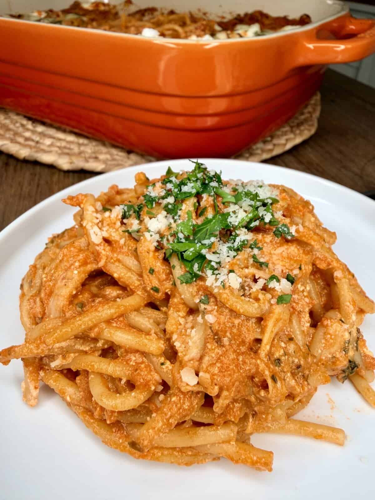A plate of baked spaghetti with red sauce, cheese, and herbs sits in the foreground, garnished with grated cheese and parsley. In the background, a large orange baking dish filled with more pasta is visible.