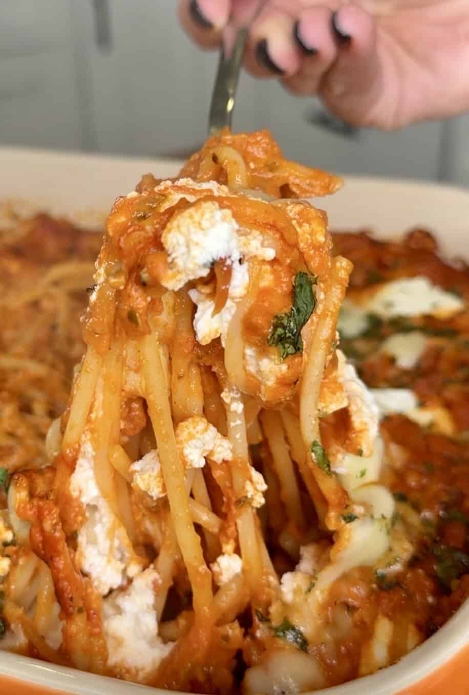 A hand holding a fork lifts a serving of baked spaghetti with tomato sauce, melted cheese, and herbs from a casserole dish.